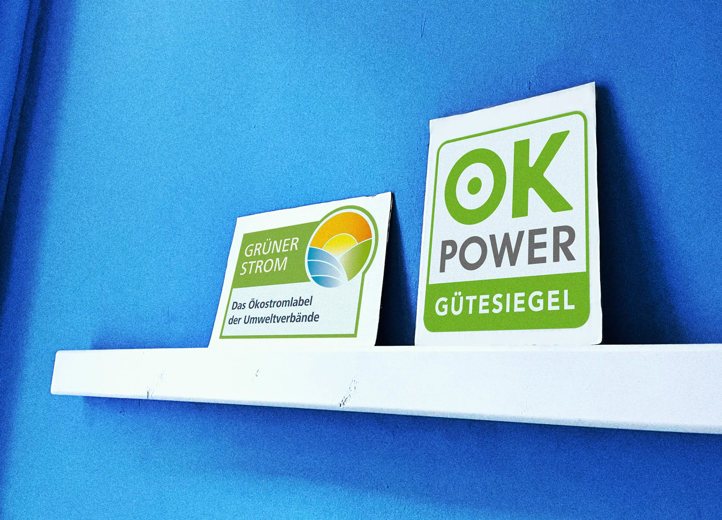 Two green energy signs, "Grüner Strom" and "OK Power Gütesiegel," stand on a white shelf against a blue wall.