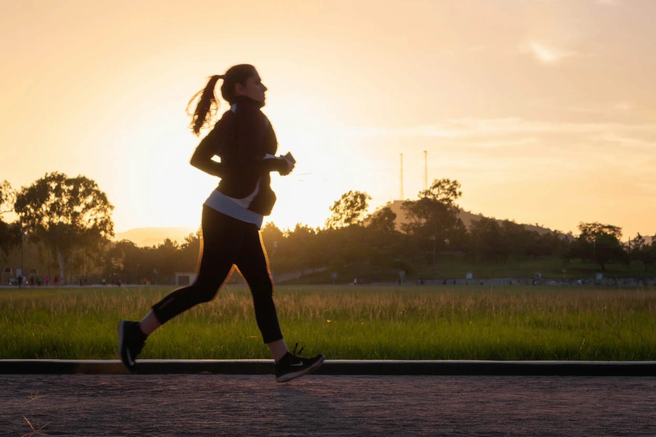 Eine Frau joggt im Sonnenuntergang neben einer Wiese und einem Hügel.