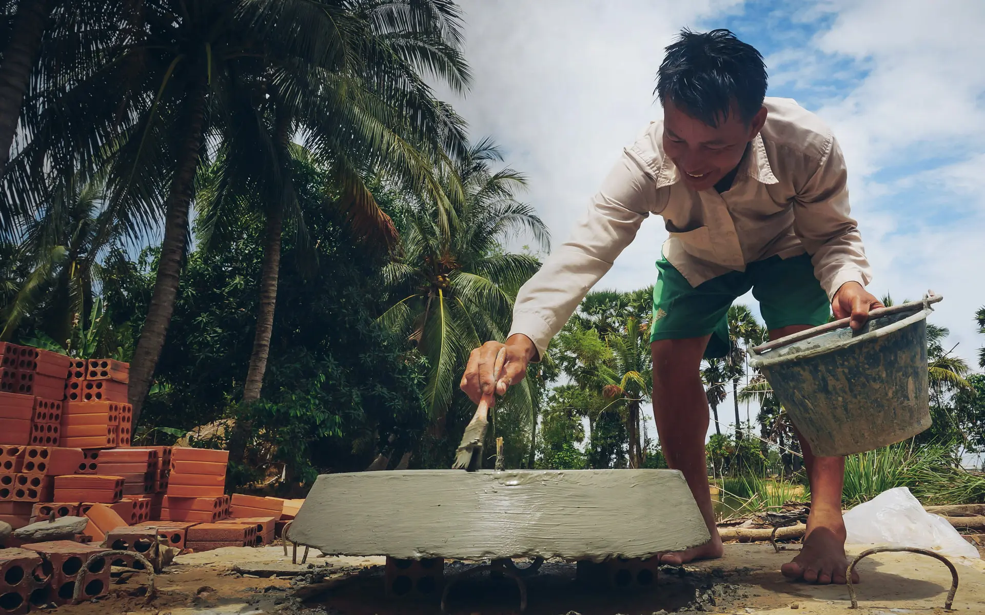 Ein Mann baut mit verschiedenen Materialien eine Biogasanlage im Boden vor Palmen und hält dabei einen Eimer in der Hand