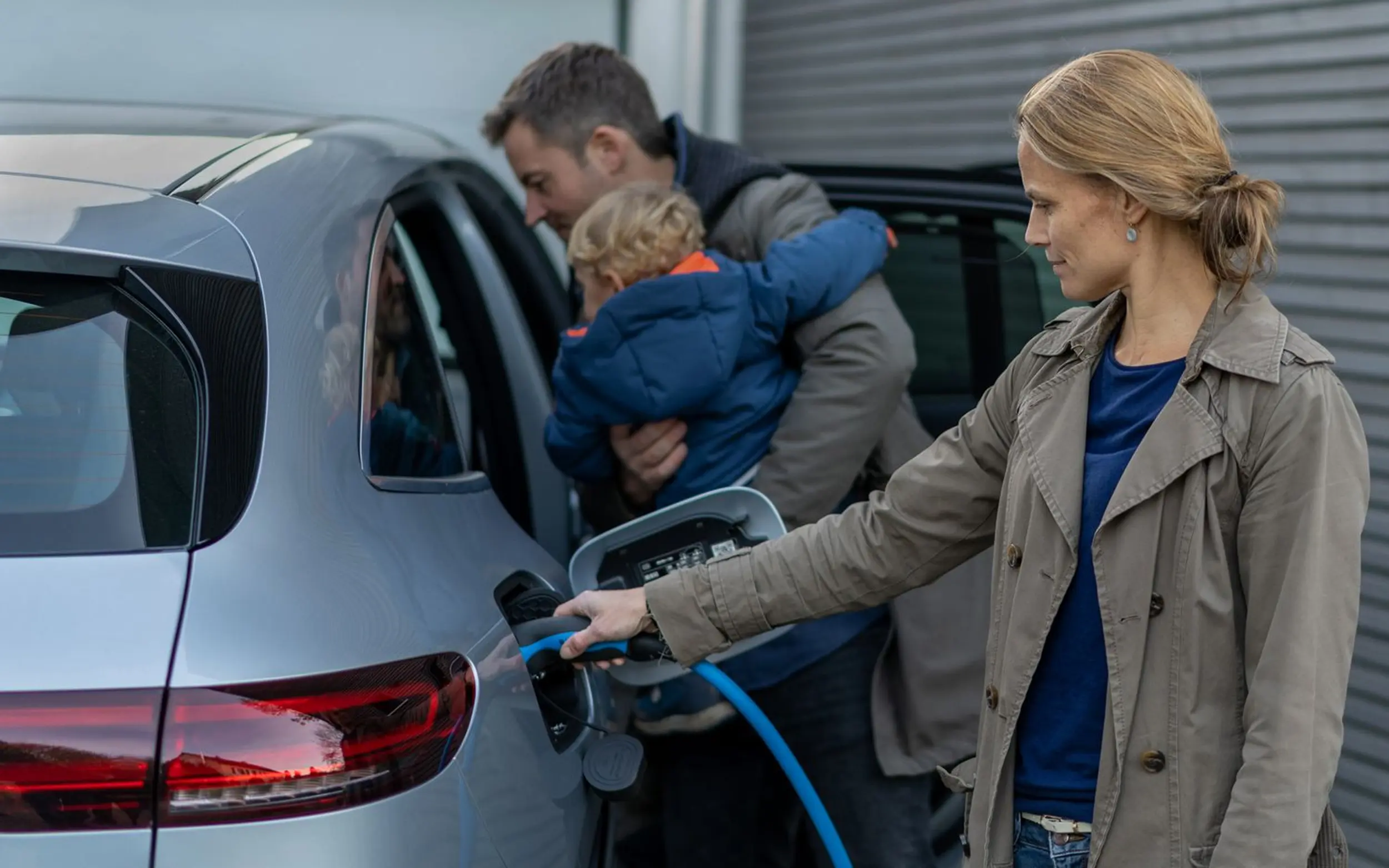 Eine Frau lädt mit einem Ladekabel das E-Auto, während der Mann das Kind ins Auto trägt