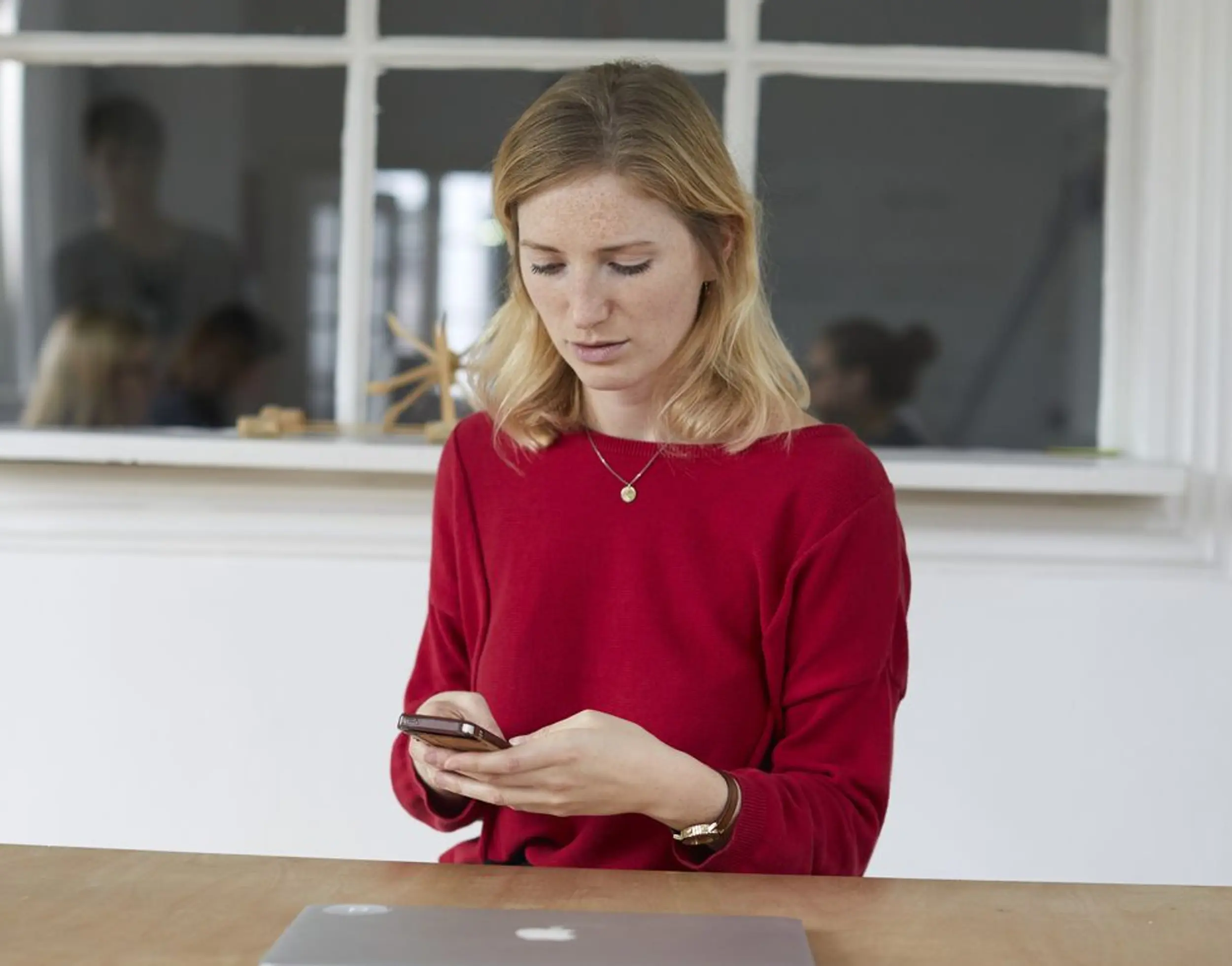 Eine junge Frau in einem roten Hemd benutzt konzentriert ihr Smartphone, während sie an einem Holztisch mit einem Laptop sitzt.