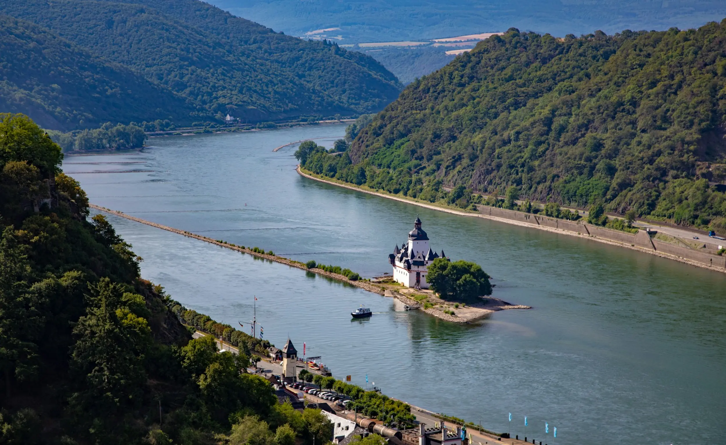 A river winds through a valley of forested hills, with a white castle on a small island in the middle of the river.