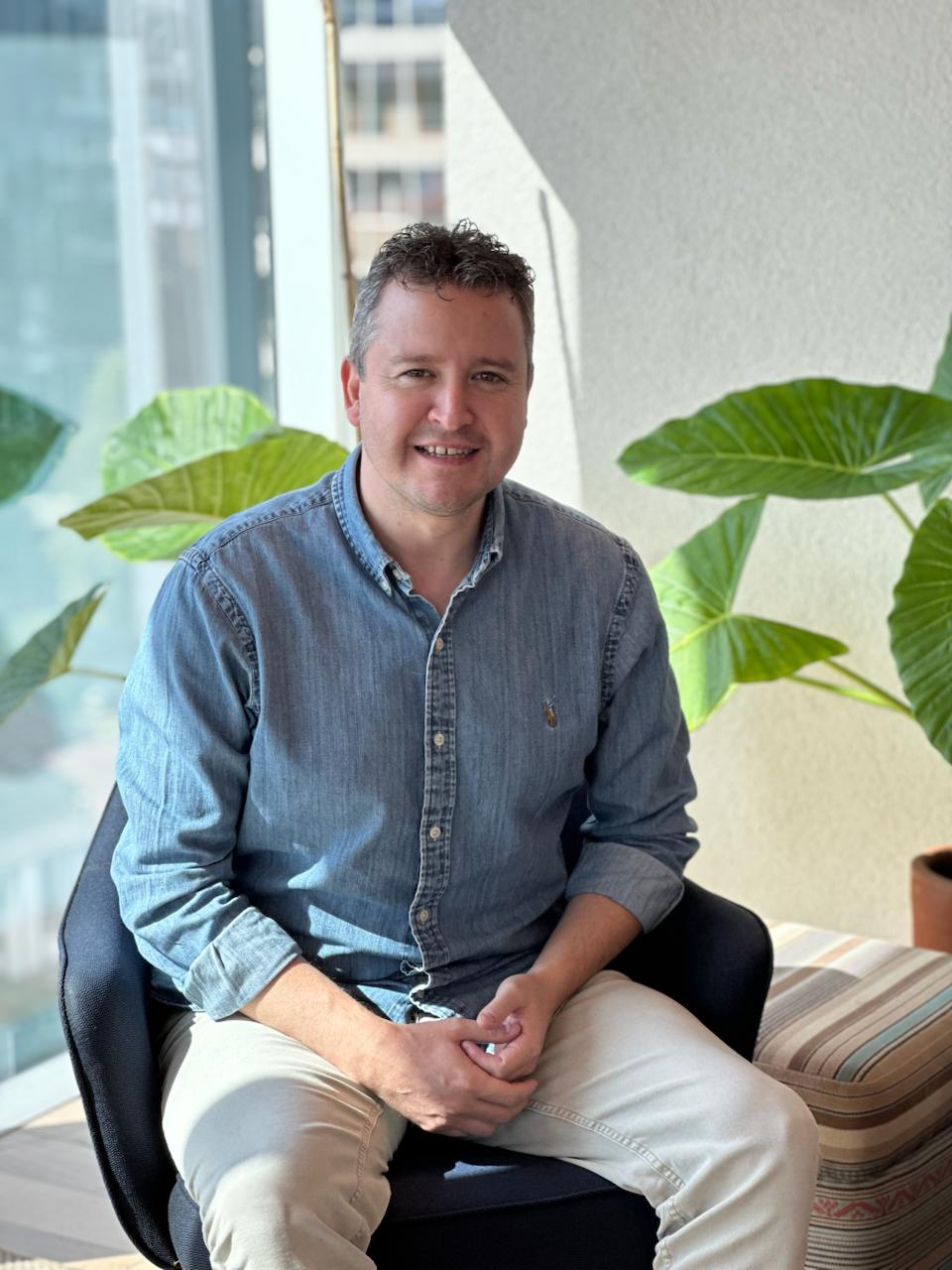A smiling man in a denim shirt sits in a modern office with large plants.