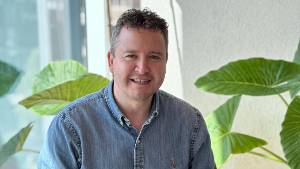 A smiling man in a denim shirt sits in a modern office with large plants.