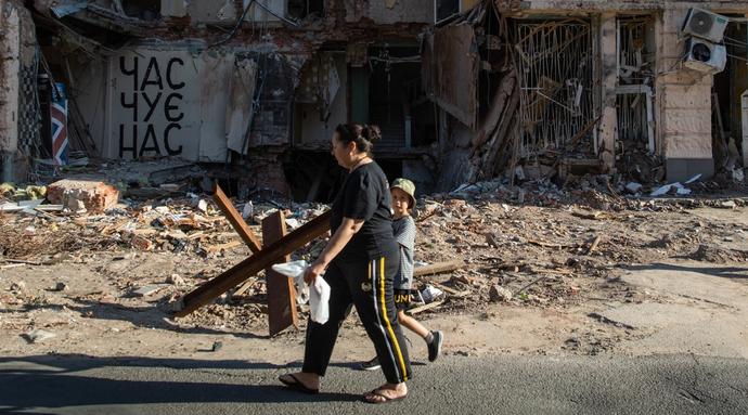 A woman walks with a child on the street. In the background a building which has been destroyed can be seen.