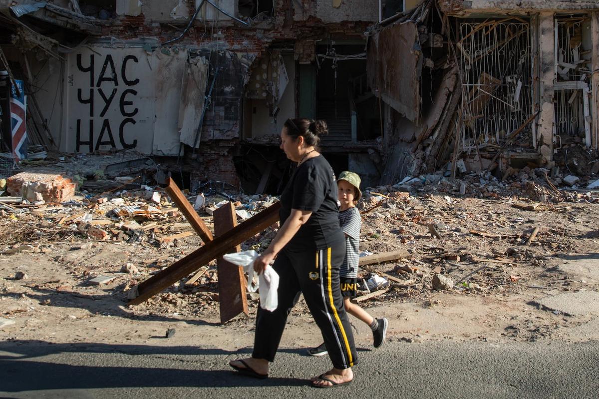 A woman walks with a child on the street. In the background a building which has been destroyed can be seen.