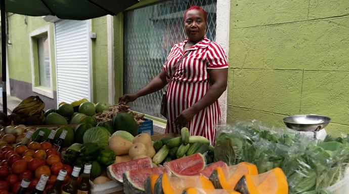 A woman stands outside behind a fruit and produce stand