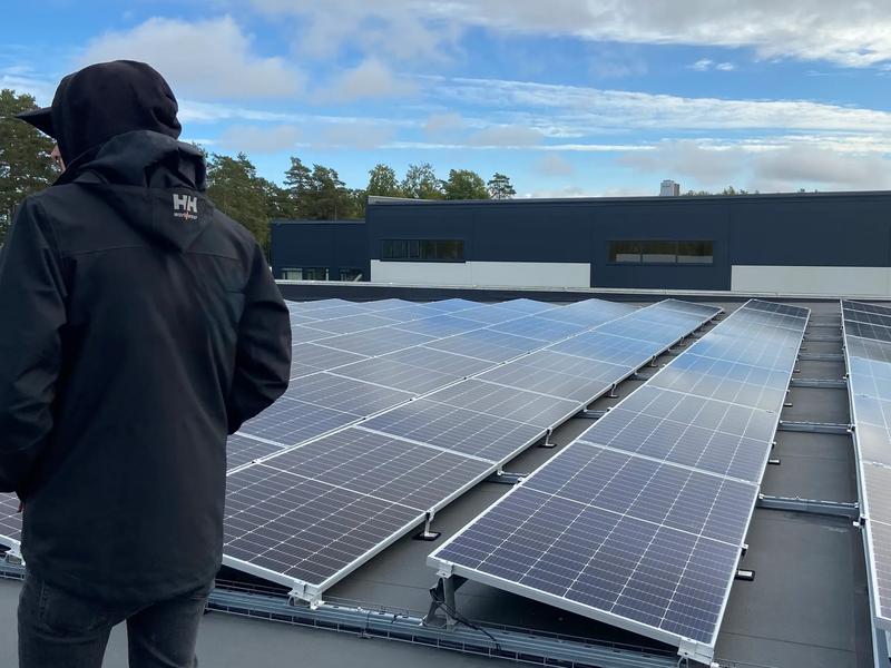 Rader av solpaneler på stativ på ett platt tak, med en företagsbyggnad i bakgrunden