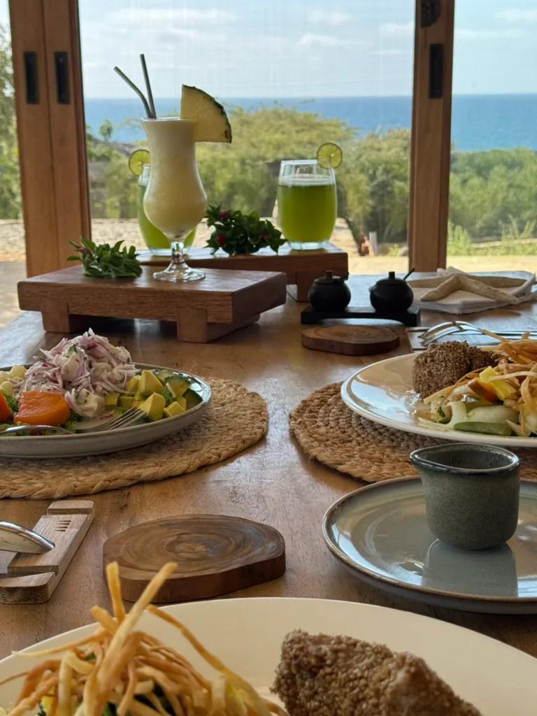 Platos de comida y dos bebidas tropicales sobre una mesa de madera con vistas al Pacífico.