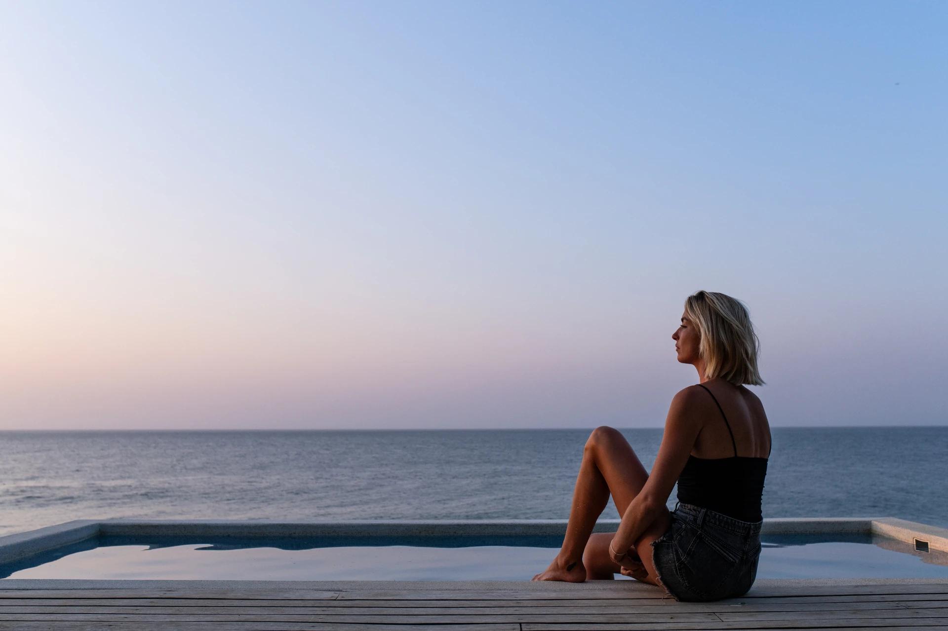 Mujer sentada junto a una piscina con vistas al Pacífico al anochecer.
