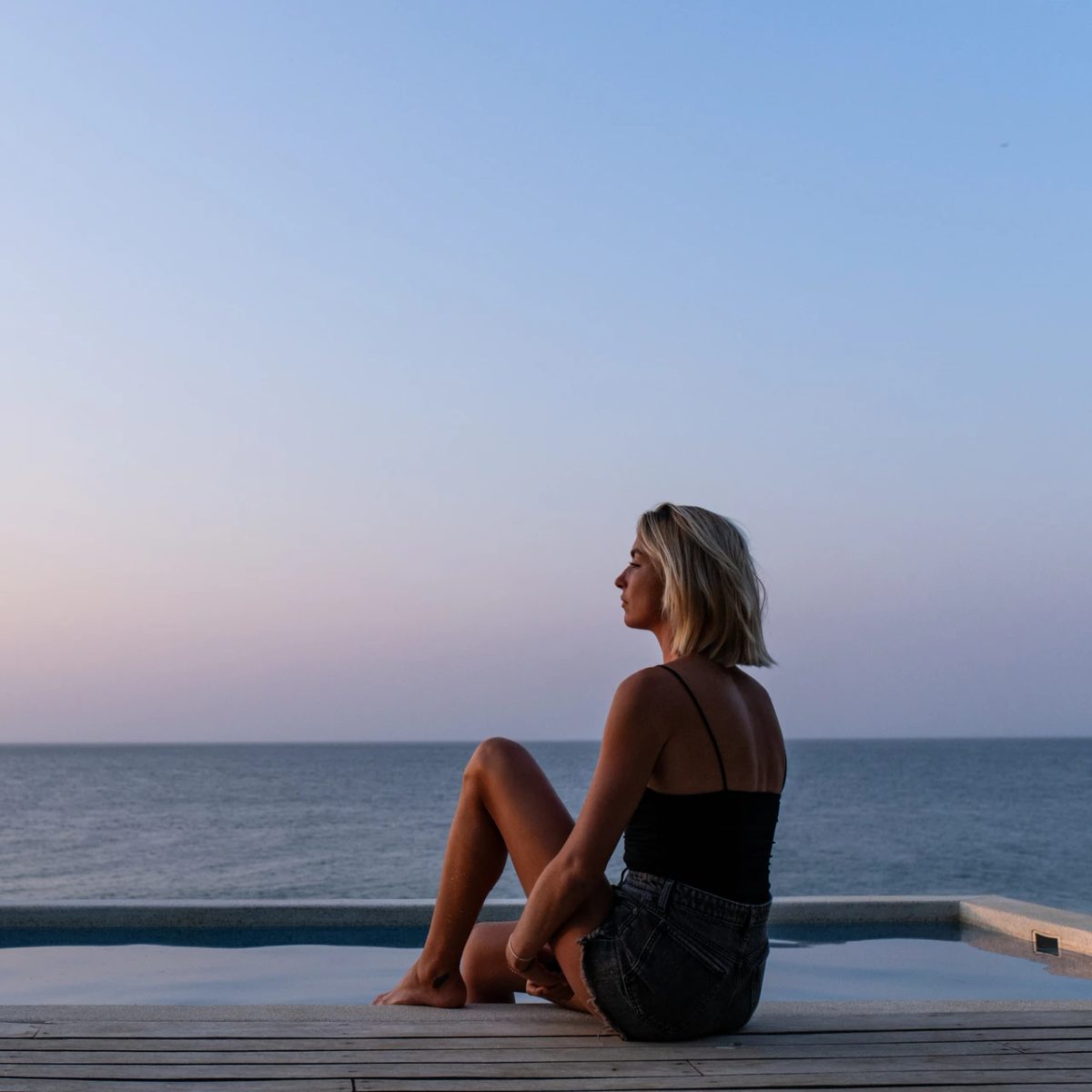 Mujer sentada junto a una piscina con vistas al Pacífico al anochecer.