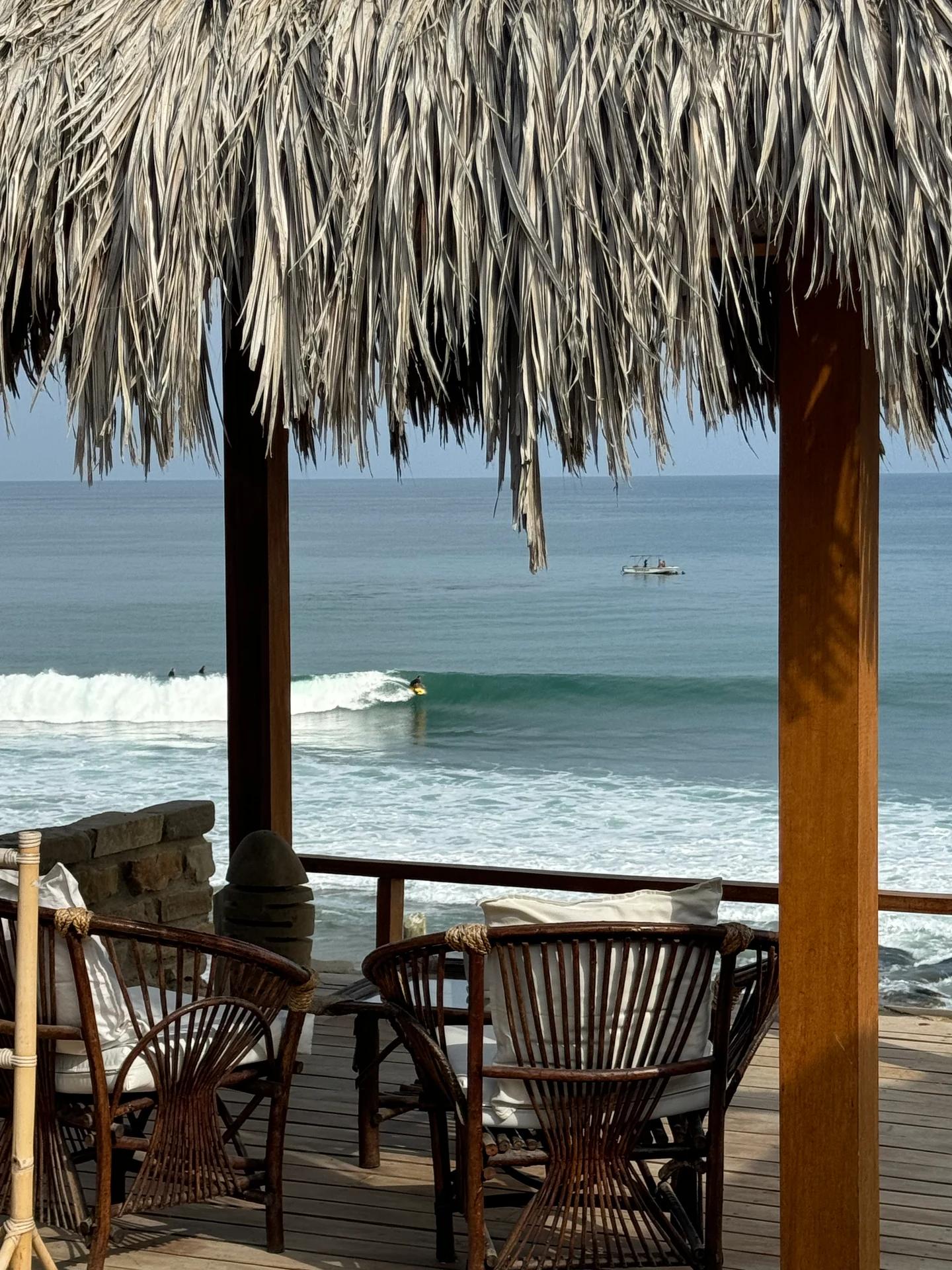 Terraza techada frente al océano con una persona surfeando una ola a la distancia.