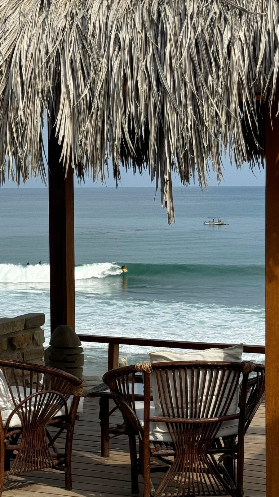 Terraza techada frente al océano con una persona surfeando una ola a la distancia.