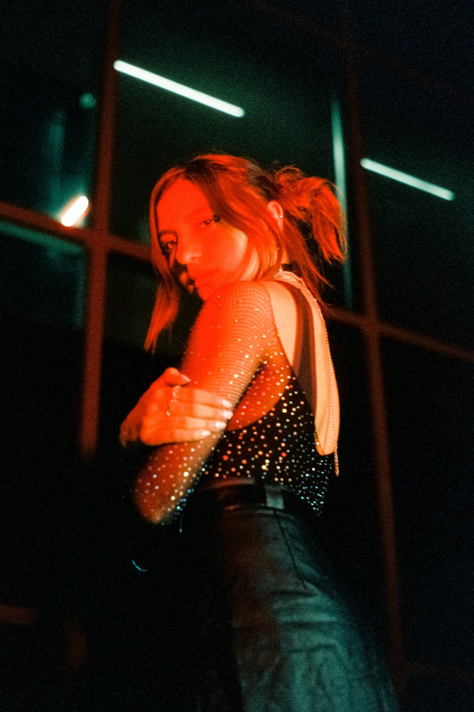 Angela Hernandez looking over her shoulder by dark windows and neon lights, photographed at night with red and green tones.
