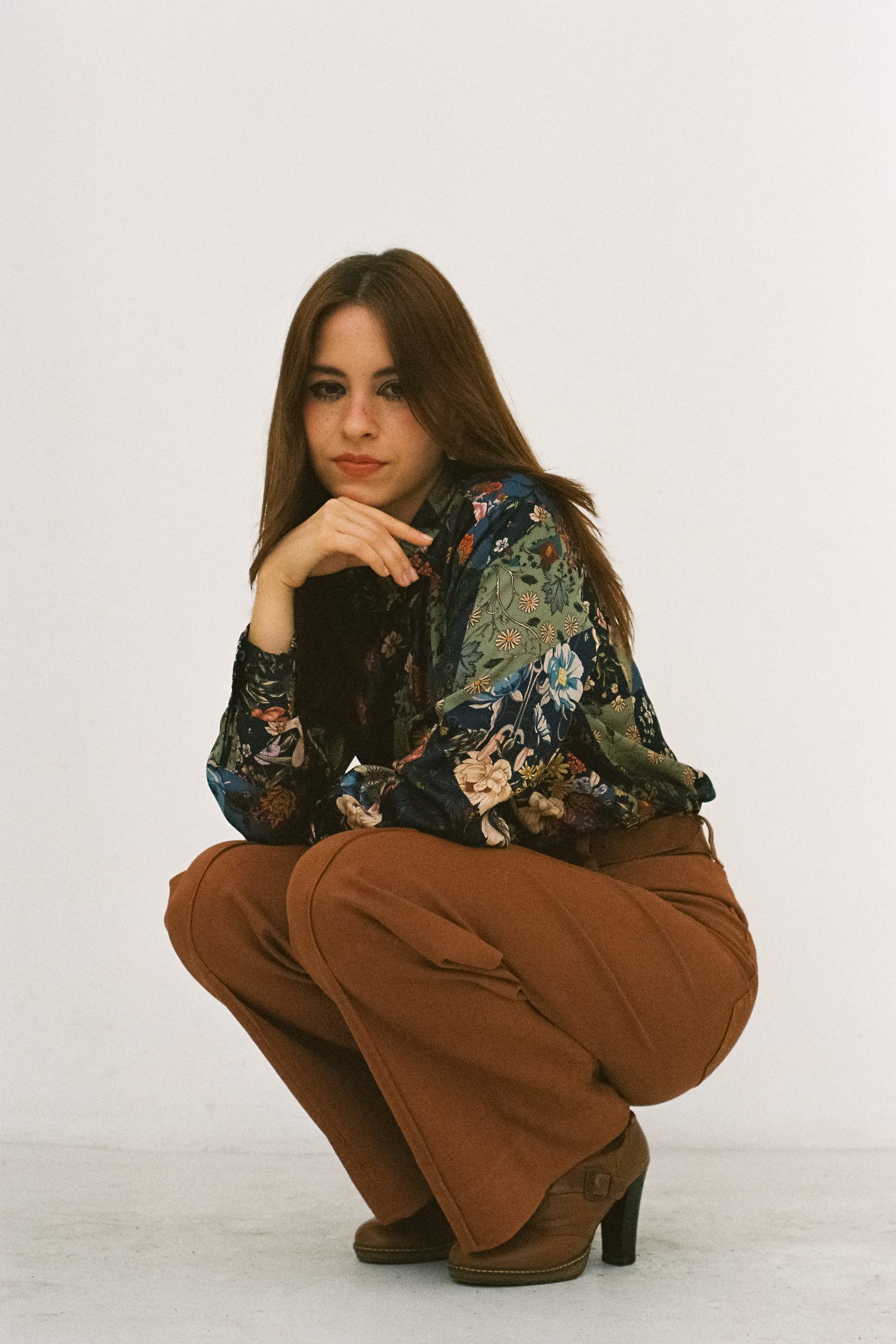Karen Cantu crouching against a white backdrop, wearing a dark floral shirt, brown flared pants, and heeled boots, with a calm pose and direct gaze.