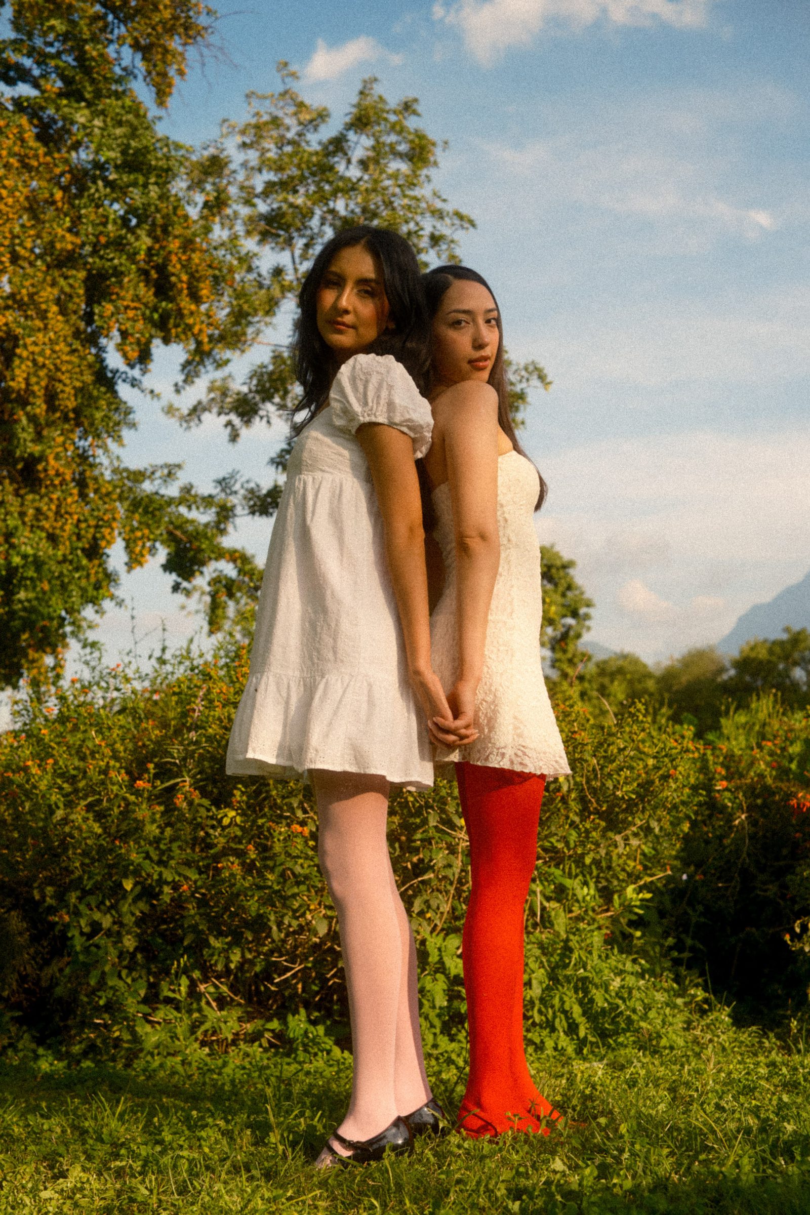 Two models in white dresses stand side by side outdoors, framed by trees and warm sunlight.