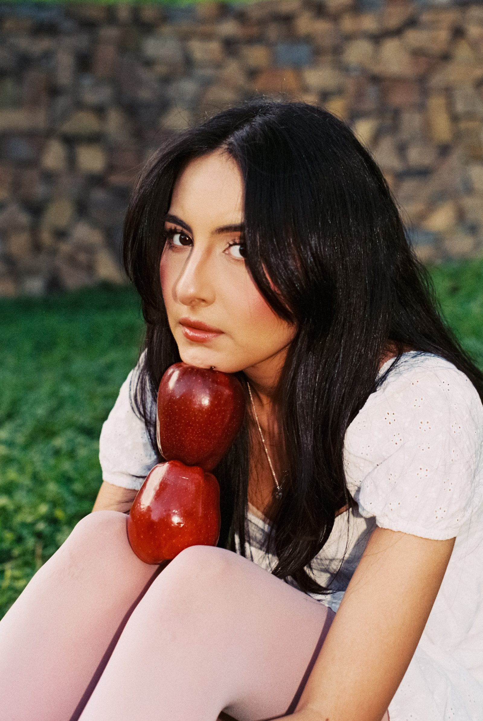 Portrait of a model in a white dress holding two red apples against her chest.