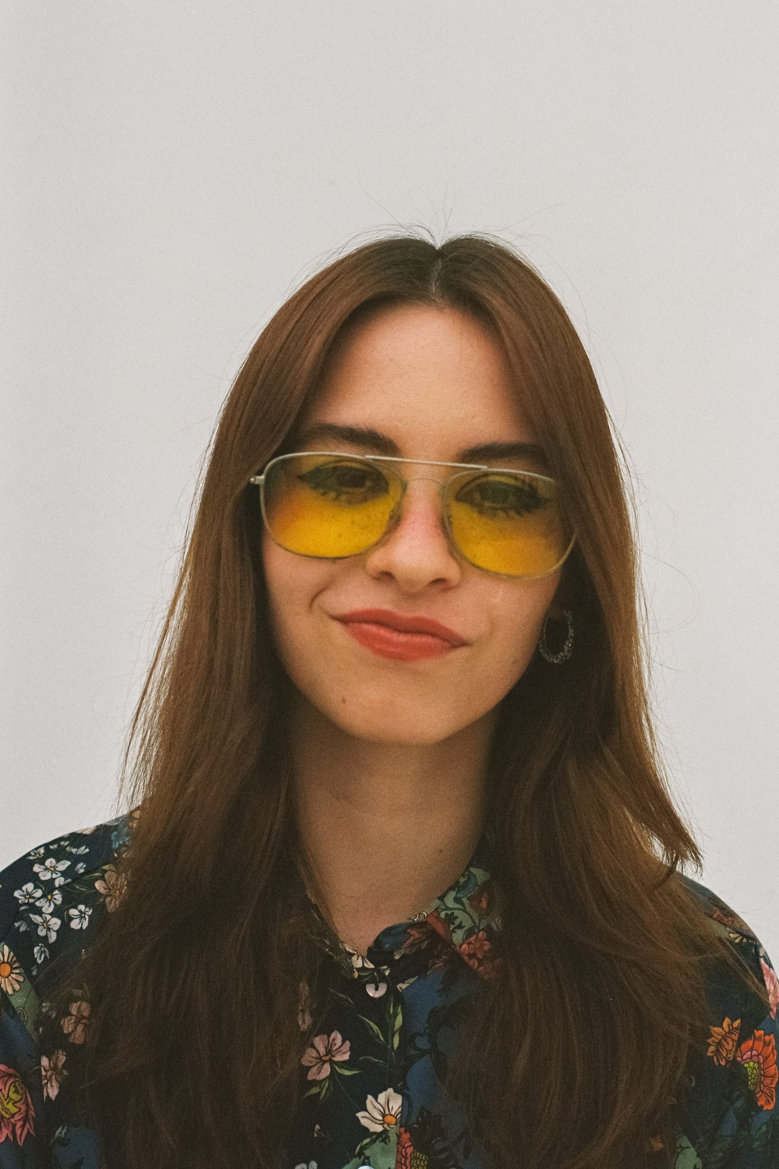 Close-up portrait of Karen Cantu wearing yellow-tinted sunglasses, bold eye makeup, and a dark floral shirt, looking at the camera with a slight smile against a minimal white background.
