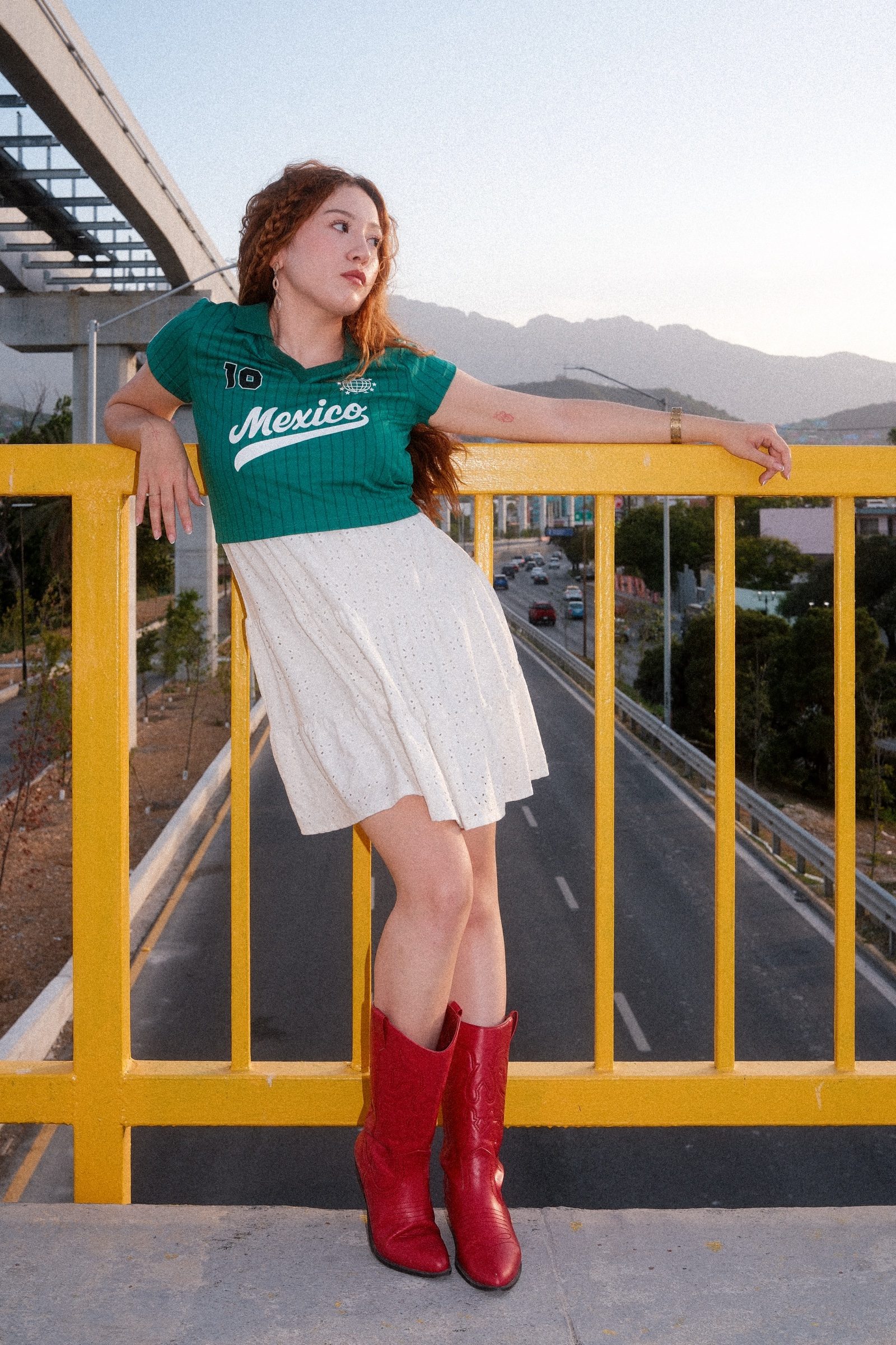 A redhaired girl dressed in Mexico's colors in a yellow bridge with the road under her and the mountains on the back