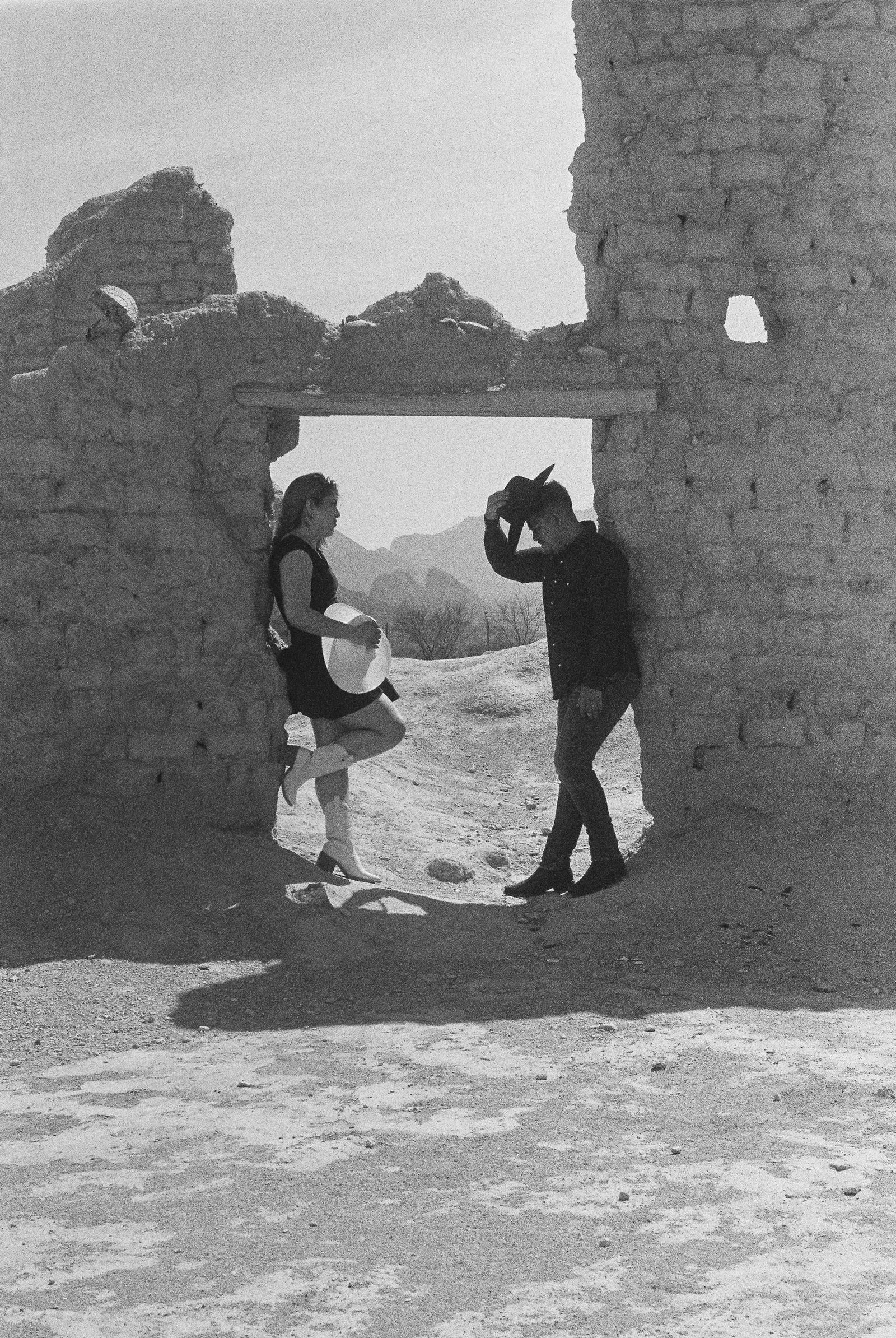 Black and white film portrait of a couple in cowboy hats standing under an adobe ruin doorway in a desert landscape.