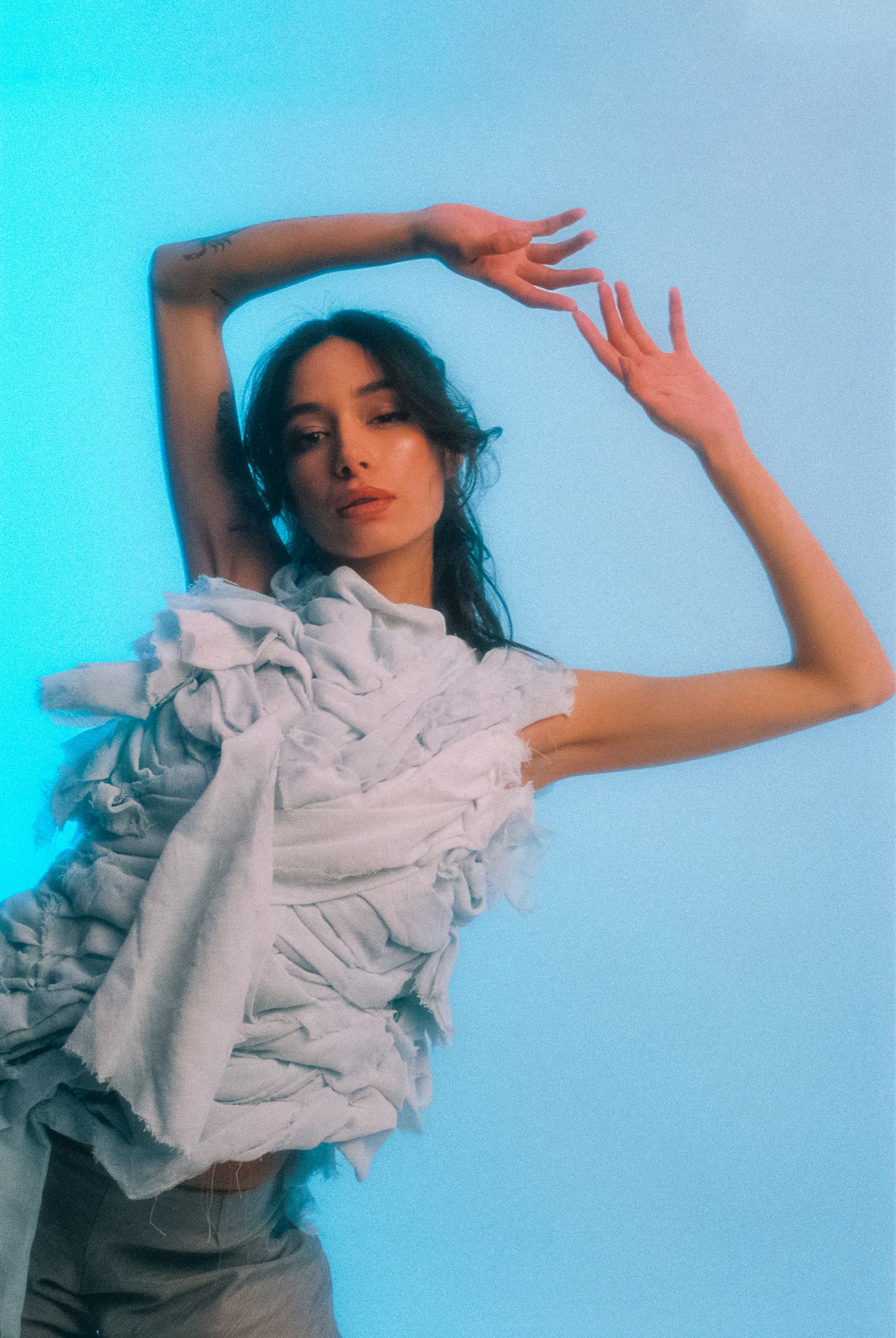 Michelle Malacara posing with raised arms in a sculptural pale fabric top, photographed on film against a cool blue studio backdrop.