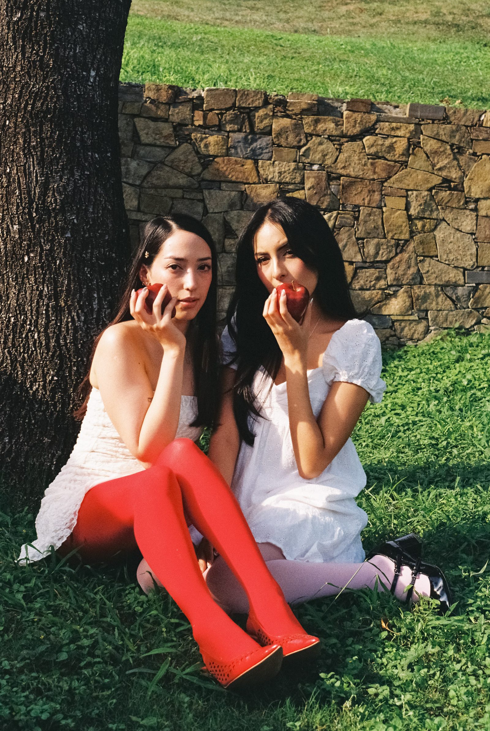 Two models sit together on the grass near a stone wall, one wearing red tights and the other a white dress.