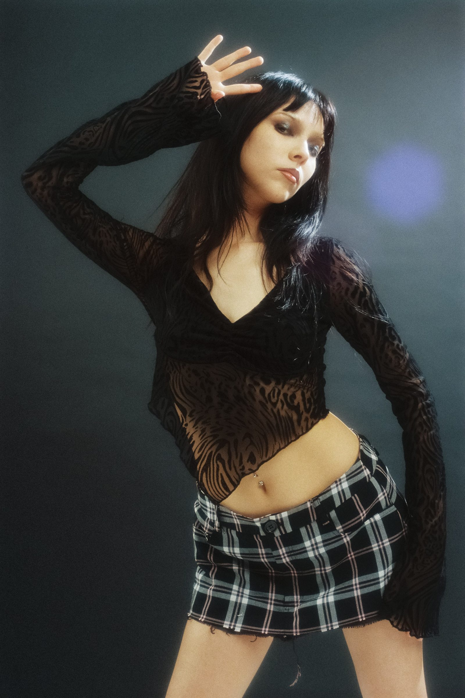 Dany Mass posing in a black sheer top and plaid skirt under soft studio light, photographed on film against a dark backdrop.