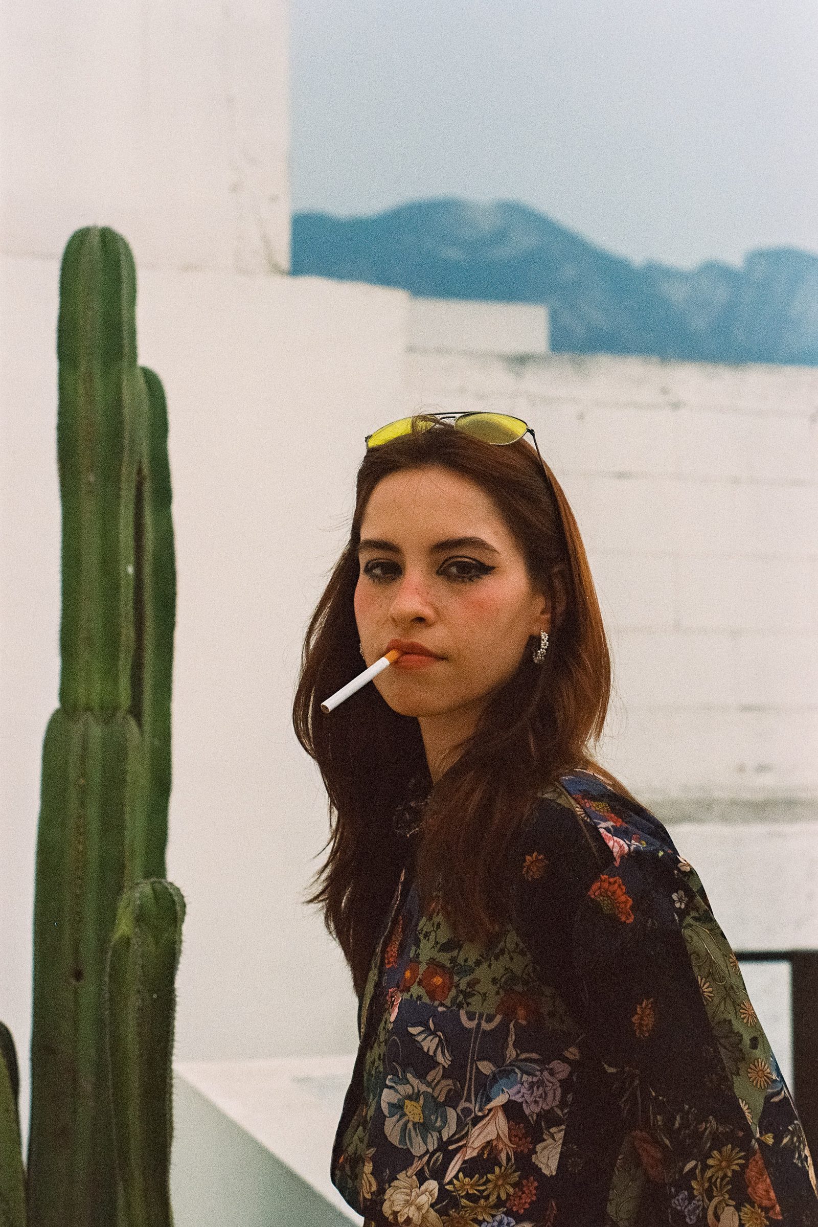Karen Cantu posing outdoors beside a cactus, wearing a floral shirt and yellow-tinted sunglasses on her head, with a cigarette in her mouth and blurred mountains in the background, captured with a vintage film aesthetic.