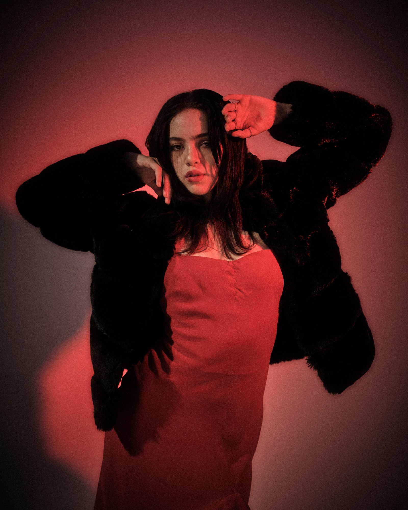 Ale Alvarado in a red dress raising her arms while wearing a black fur coat, photographed against a red-lit studio backdrop.