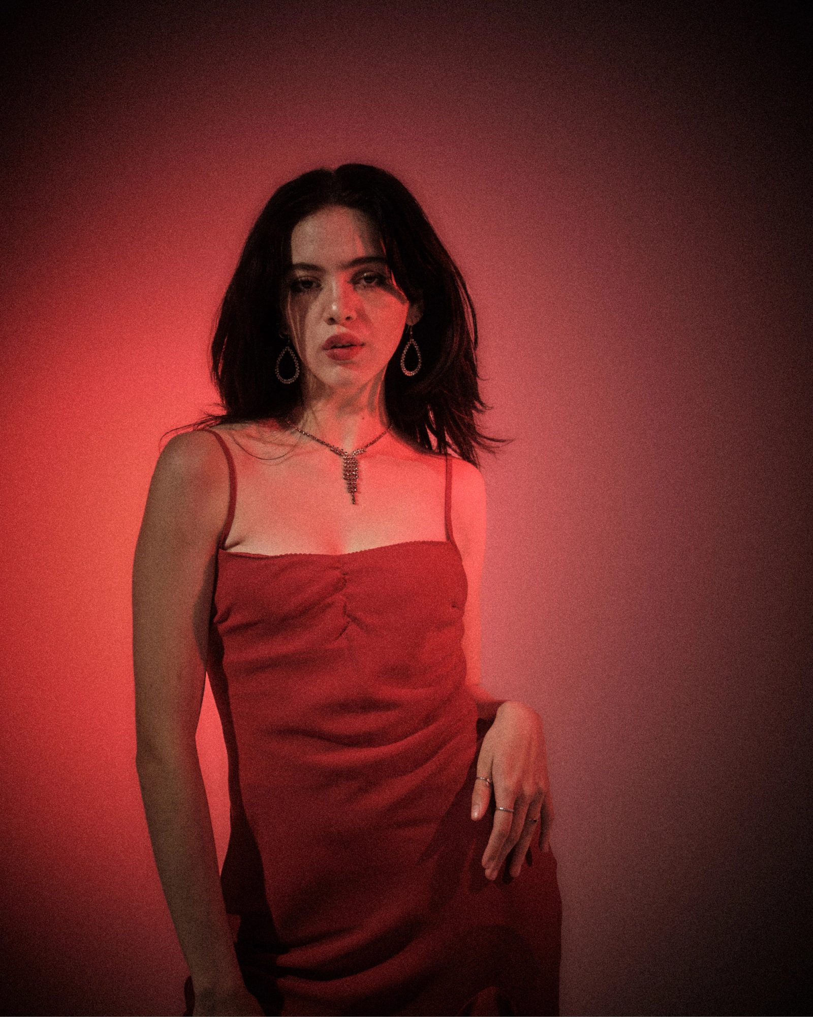 Ale Alvarado in a red dress and rhinestone necklace, photographed in red studio light with a direct femme fatale gaze.