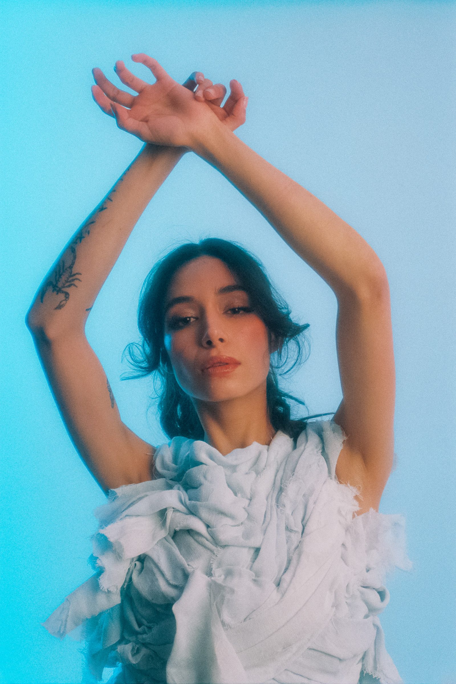 Michelle Malacara posing with crossed raised arms in a sculptural pale fabric top, photographed on film with cool blue studio lighting.