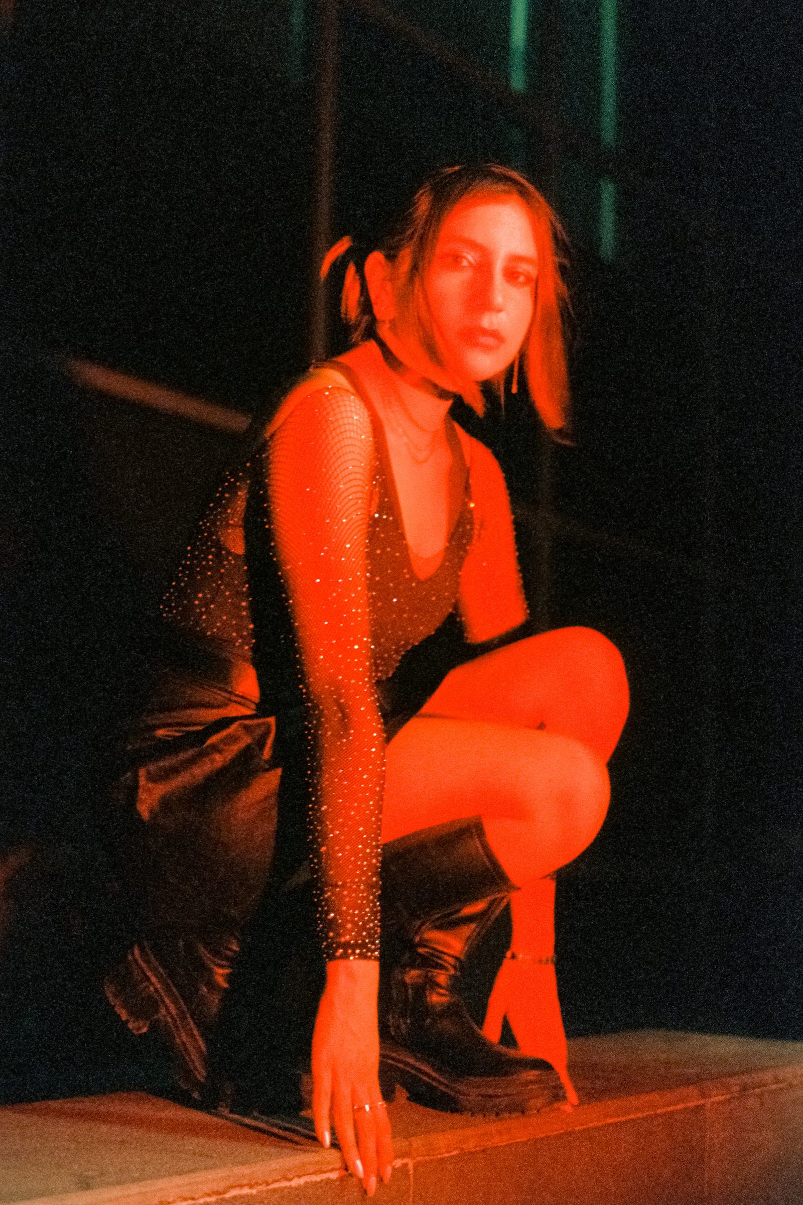Angela Hernandez crouching on a curb in a black punk outfit, photographed at night under red light with deep shadows.