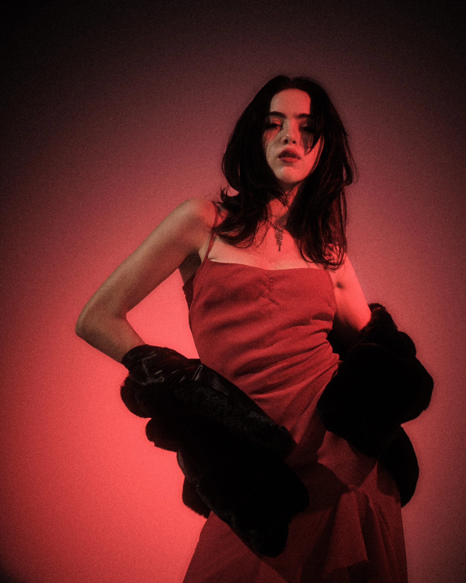 Ale Alvarado posing in a red dress with a black fur coat draped at her waist, photographed under red studio light.