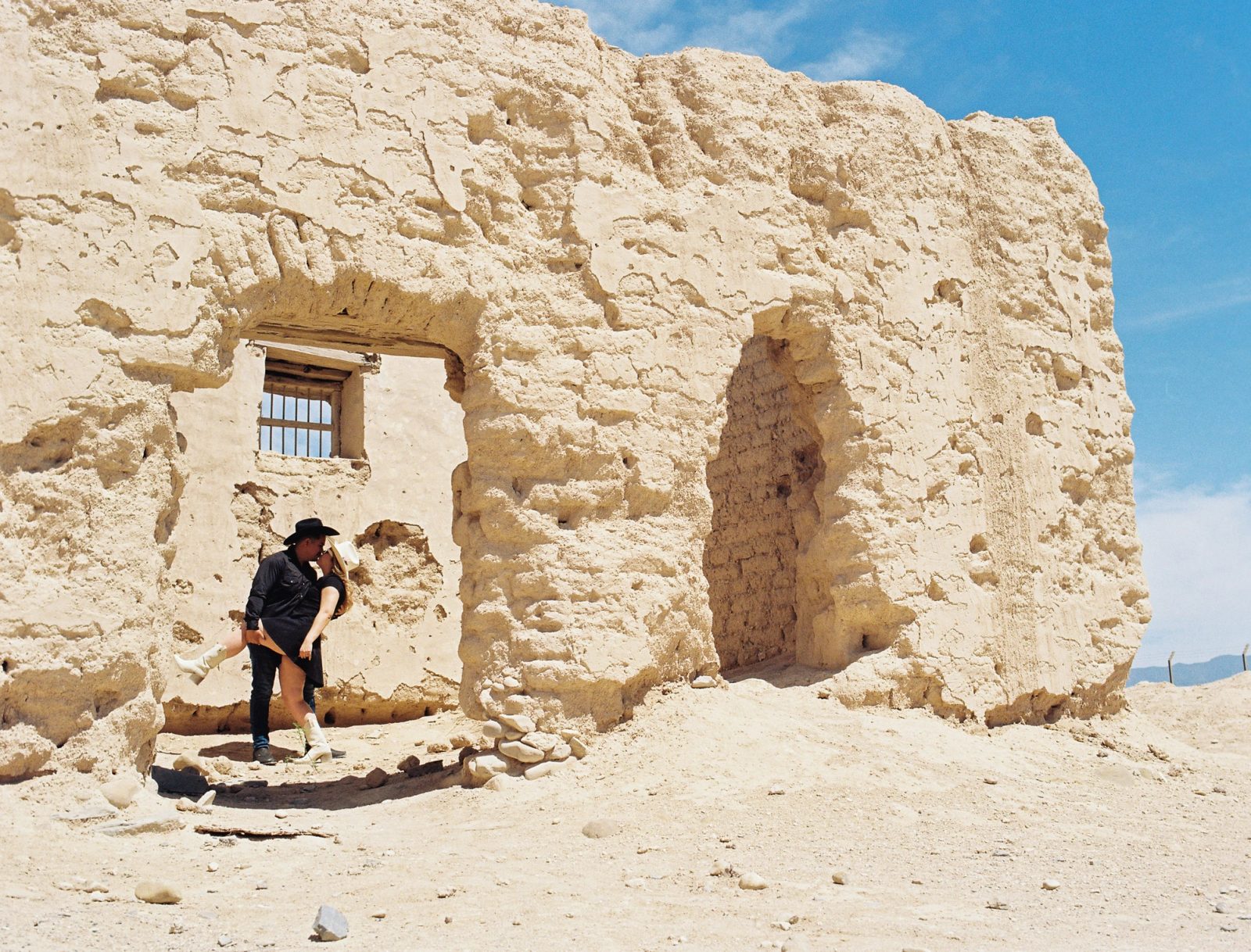 Couple kissing under weathered adobe ruins, dressed in western styling and photographed on film in bright desert light.