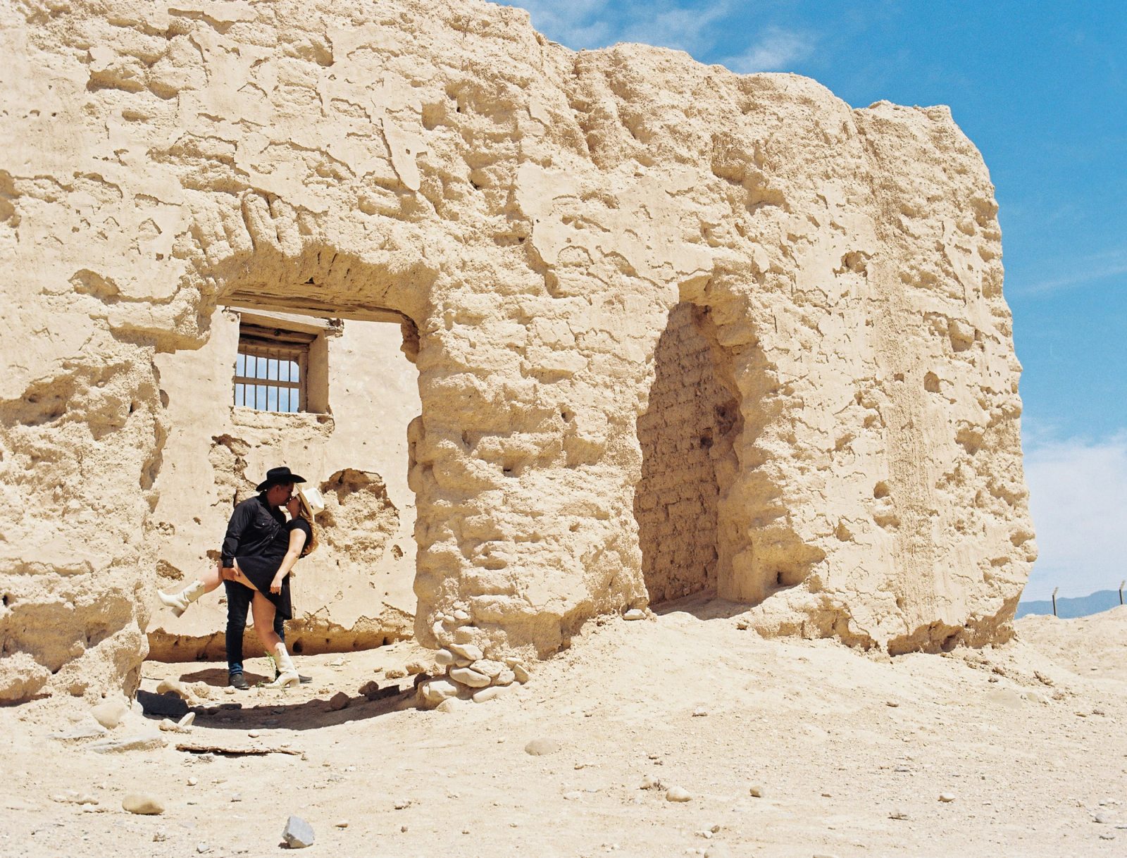 Couple kissing under weathered adobe ruins, dressed in western styling and photographed on film in bright desert light.
