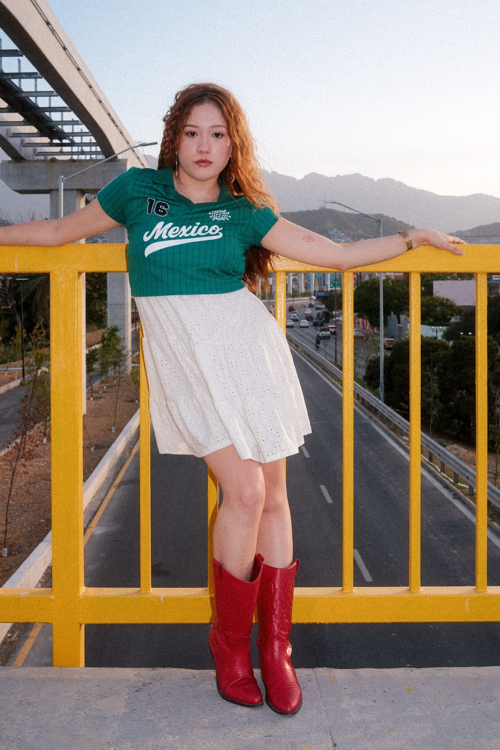A redhaired girl dressed in Mexico's colors in a yellow bridge with the road under her and the mountains on the back