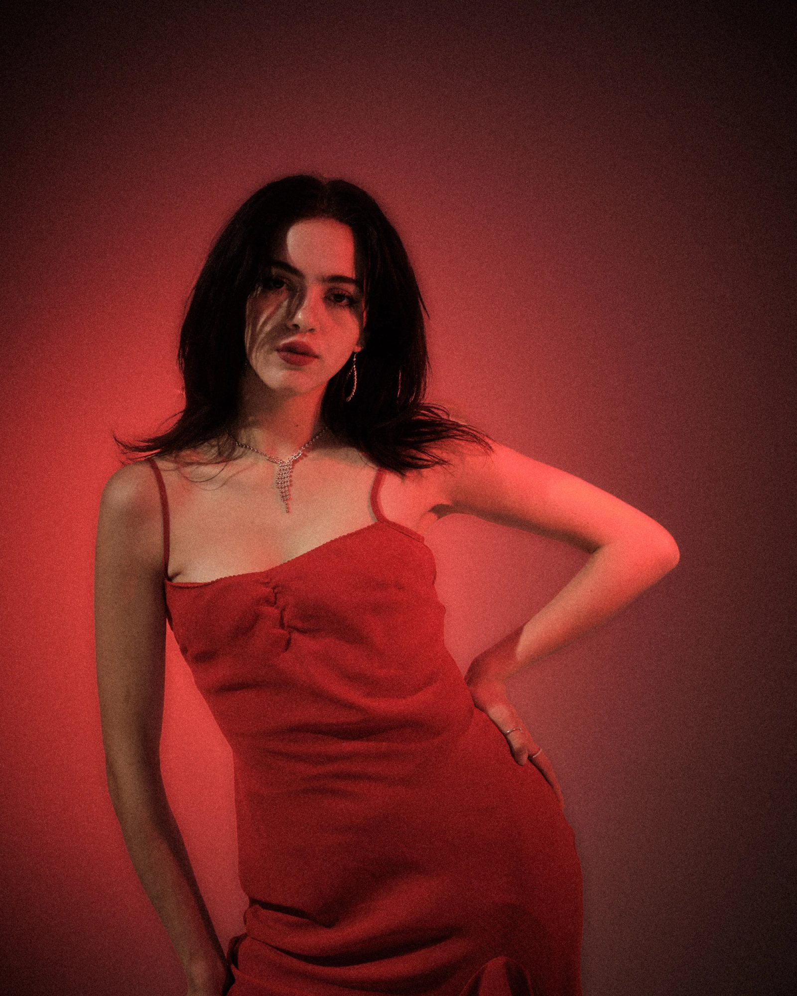 Ale Alvarado posing in a red dress against a red-lit studio backdrop, wearing drop earrings and a rhinestone necklace.