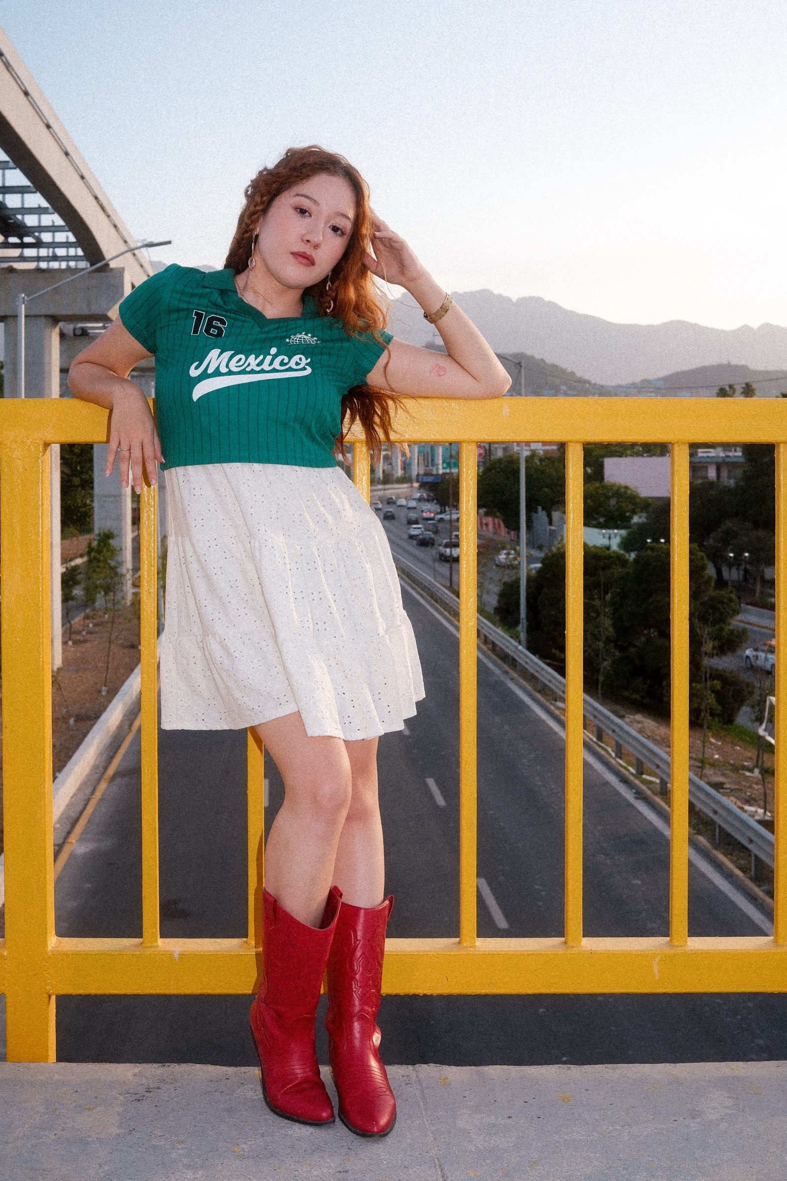 A redhaired girl dressed in Mexico's colors in a yellow bridge with the road under her and the mountains on the back