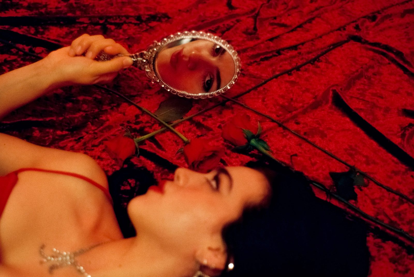 Ale Alvarado reflected in a handheld mirror while lying among roses on red velvet, photographed on film in a red studio setting.