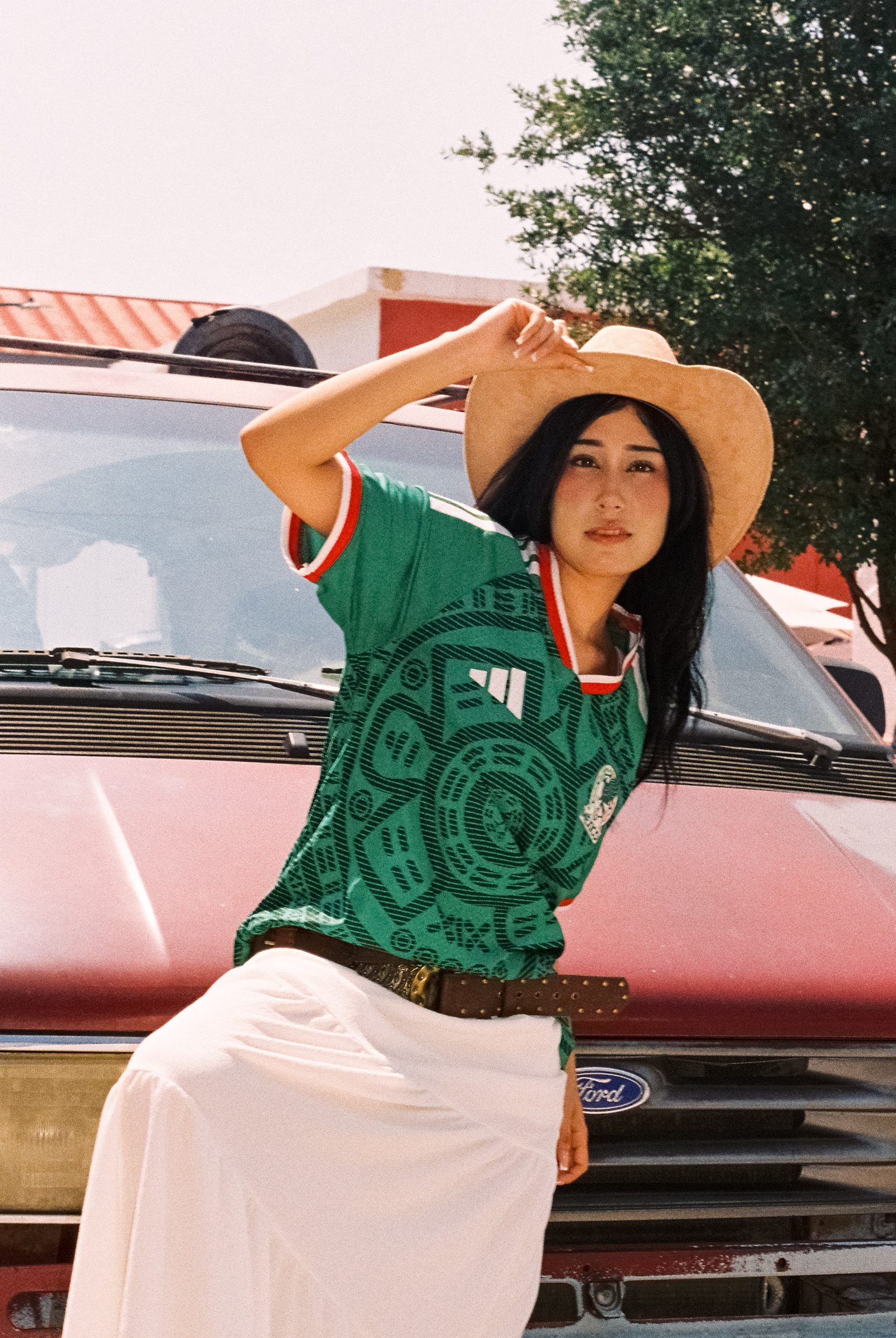 Liz con sombrero y el jersey de la Selección Mexicana con una mano en el sombrero mientras se recarga en el vehículo