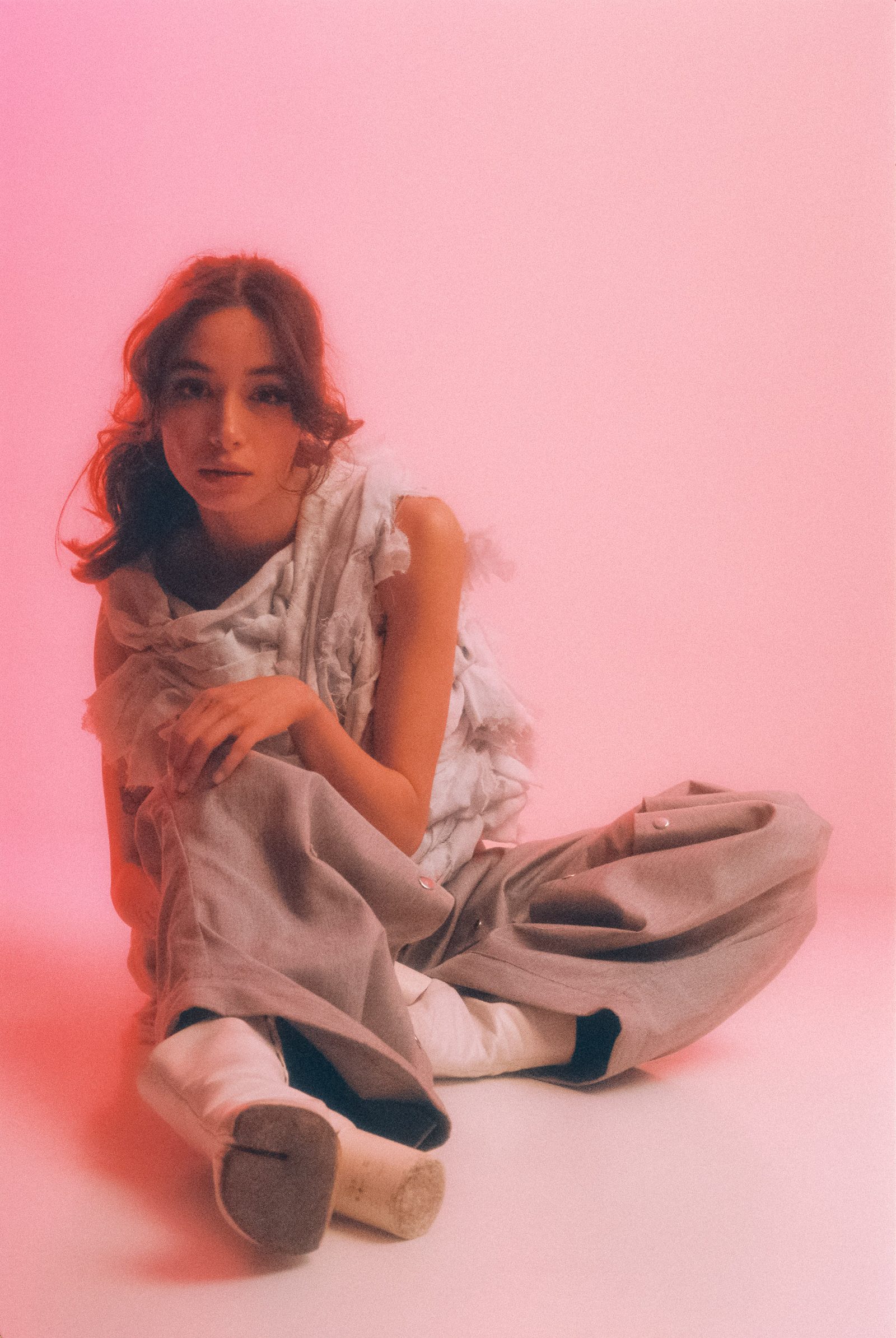 Michelle Malacara seated in gray pants and a sculptural pale fabric top against a soft pink studio backdrop, photographed on film.