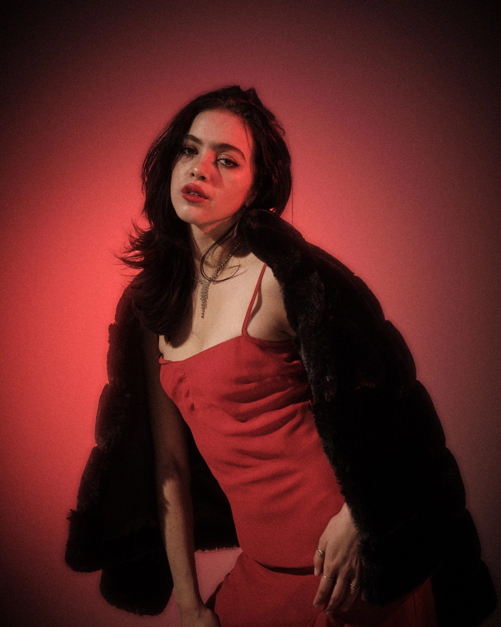 Ale Alvarado leaning forward in a red dress and black fur coat, photographed in dramatic red studio lighting.