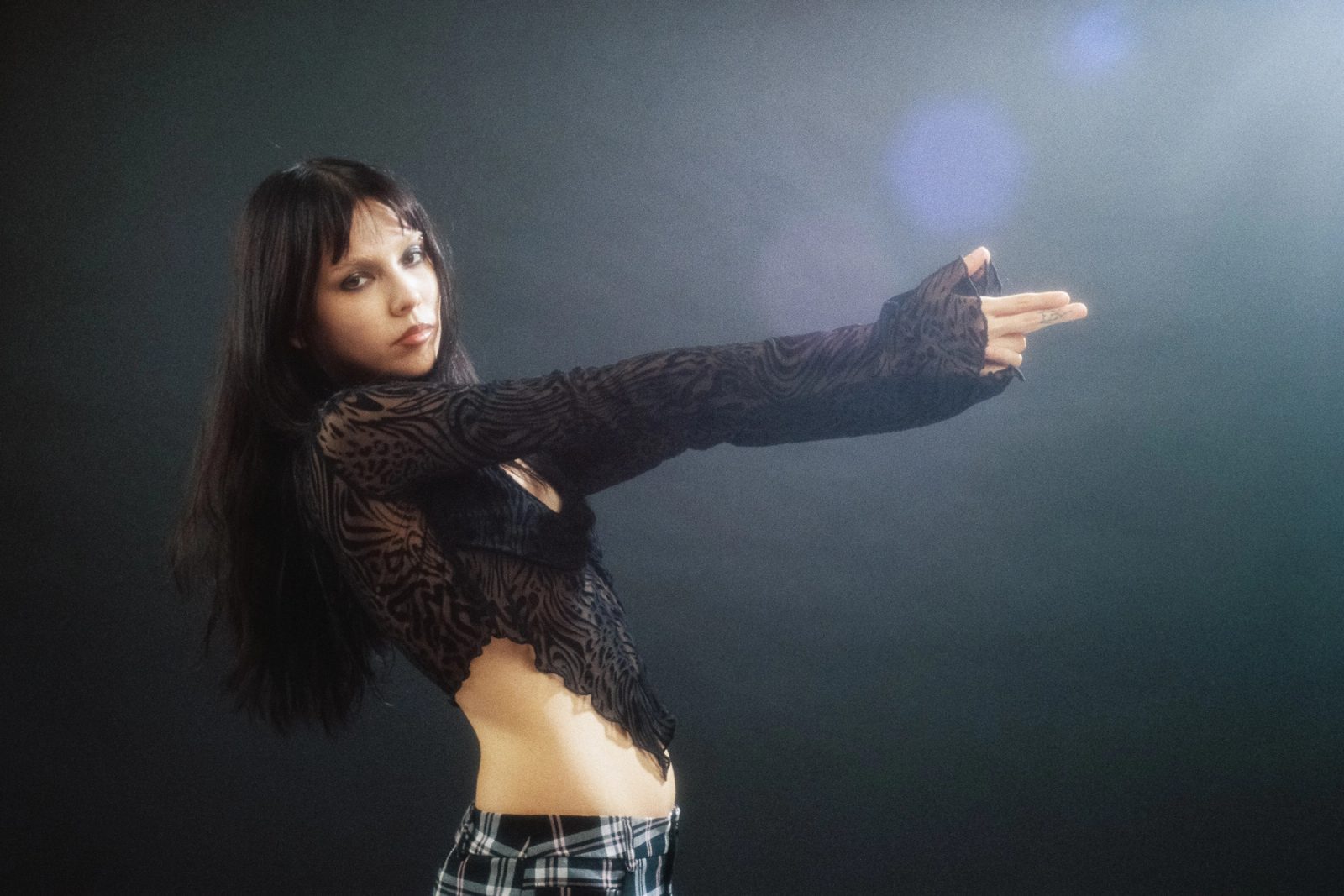 Dany Mass extending her arms in a black sheer top, photographed on film against a dark studio backdrop with soft lens flare.