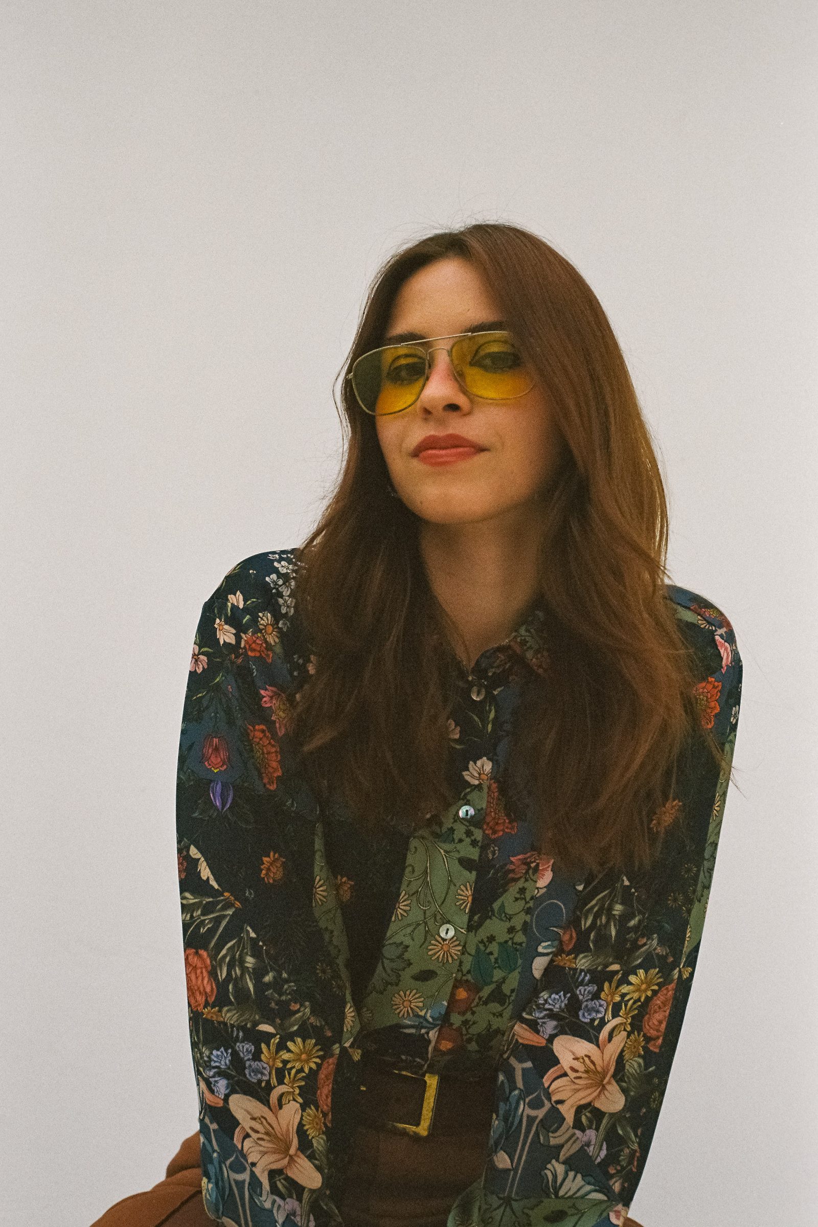 Karen Cantu posing against a white backdrop, wearing yellow-tinted sunglasses, a dark floral shirt, and brown pants, with a calm expression and a retro editorial mood.