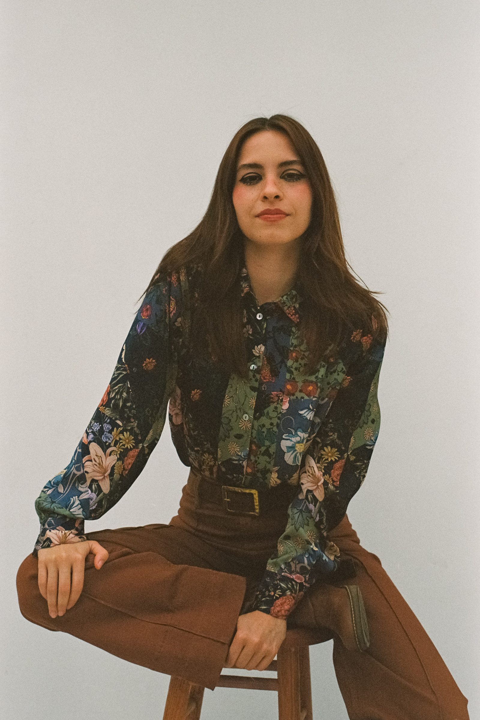 Editorial portrait of Karen Cantu sitting on a wooden stool, wearing a dark floral shirt, high-waisted brown pants, and a black belt, posed against a white studio backdrop with a vintage film aesthetic.