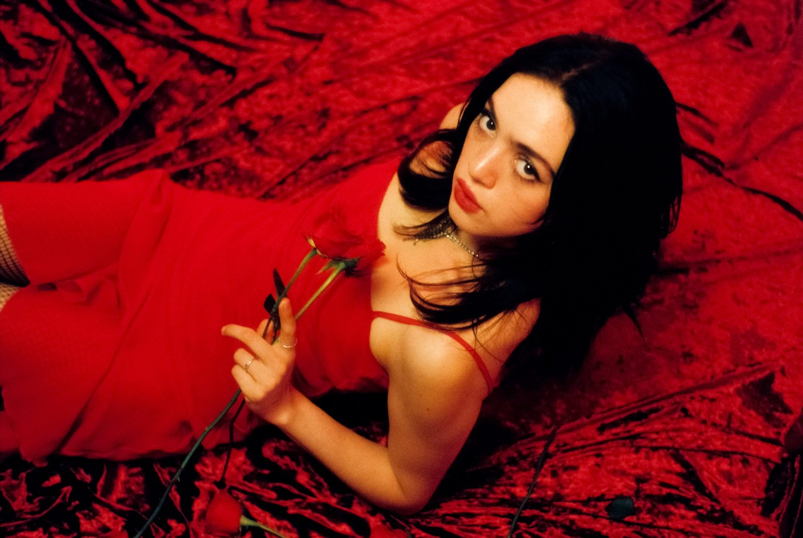 Ale Alvarado holding a red rose while lying on red velvet, photographed on film in an intimate red-toned portrait.