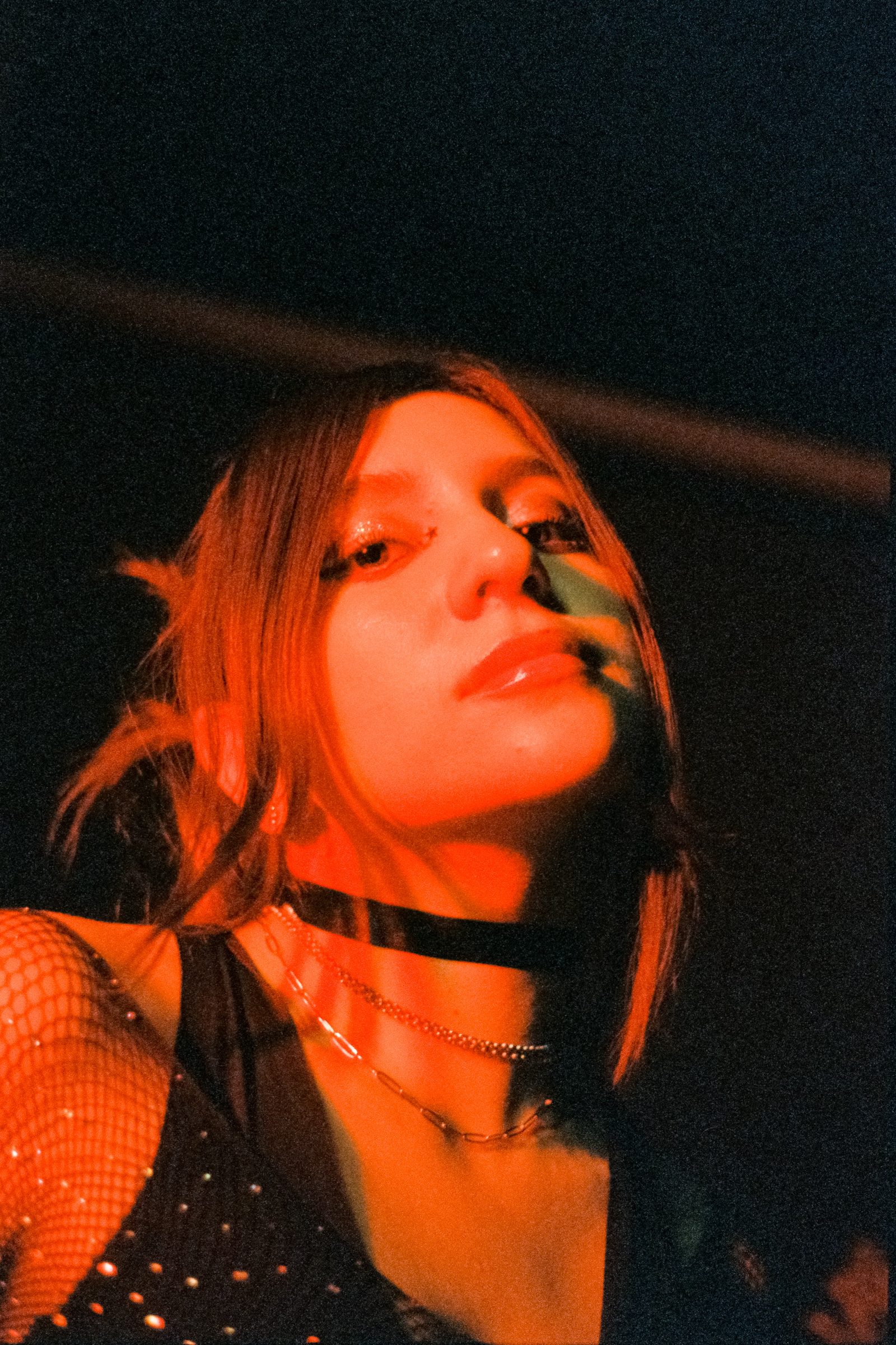Close-up portrait of Angela Hernandez under intense red light, wearing a black choker and layered necklaces at night.