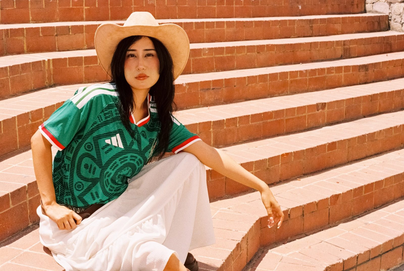 Liz con sombrero y el jersey de la Selección Mexicana sentada en unas escaleras de ladrillo