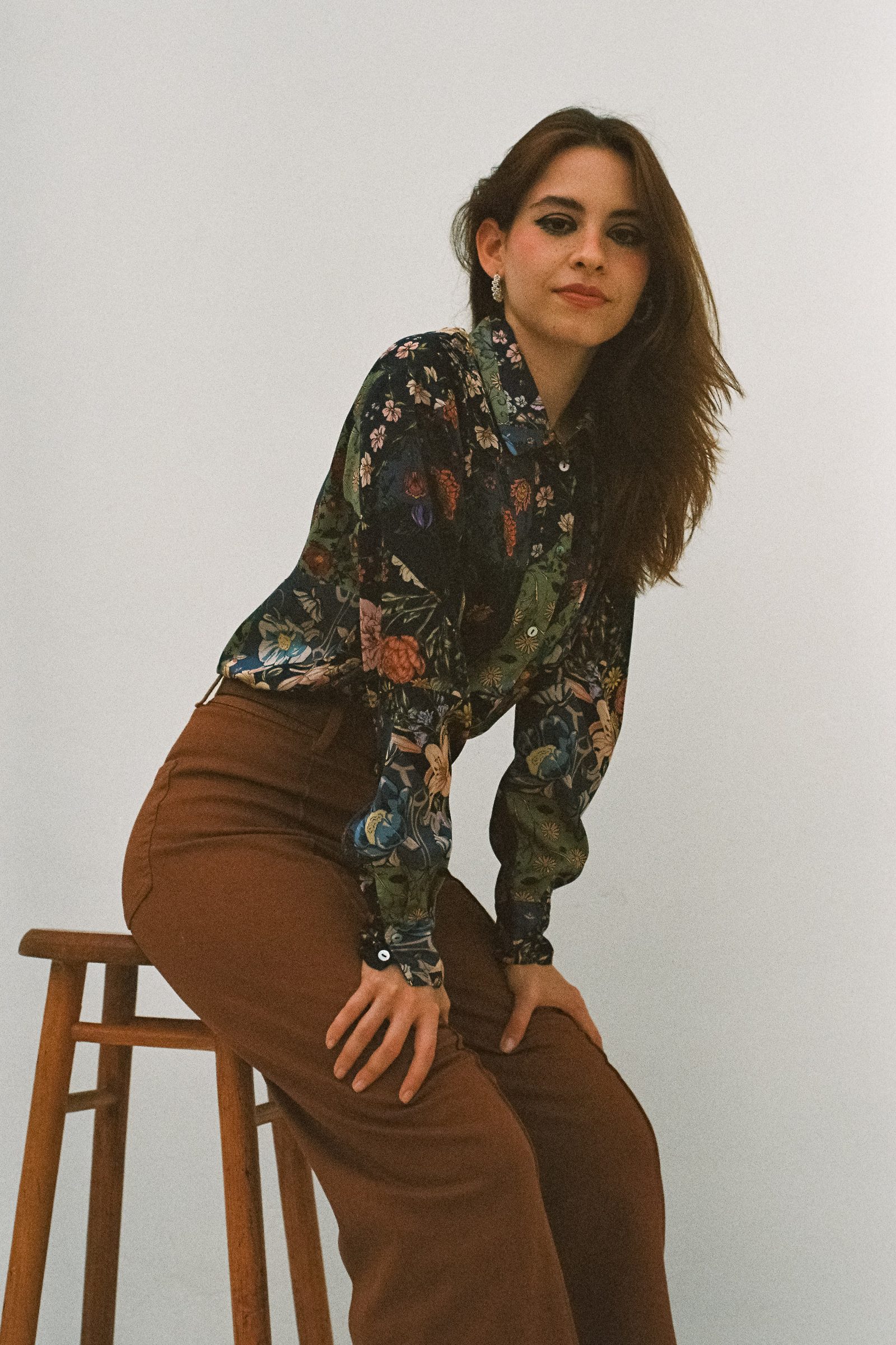 Karen Cantu sitting sideways on a wooden stool, wearing a floral shirt and brown pants, leaning toward the camera in an editorial pose against a white studio backdrop.
