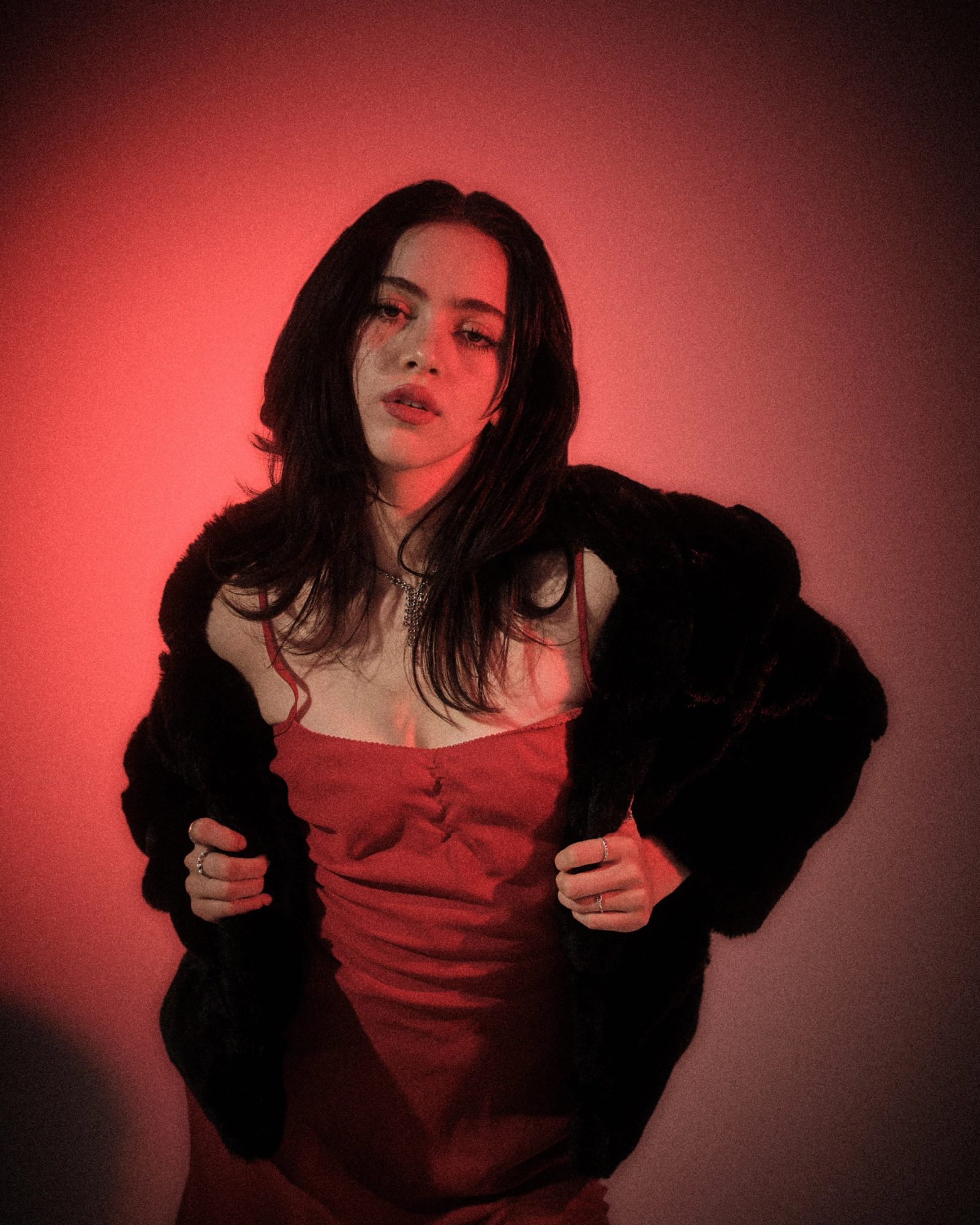 Ale Alvarado in a red dress and black fur coat, photographed in red studio light with a sharp femme fatale mood.